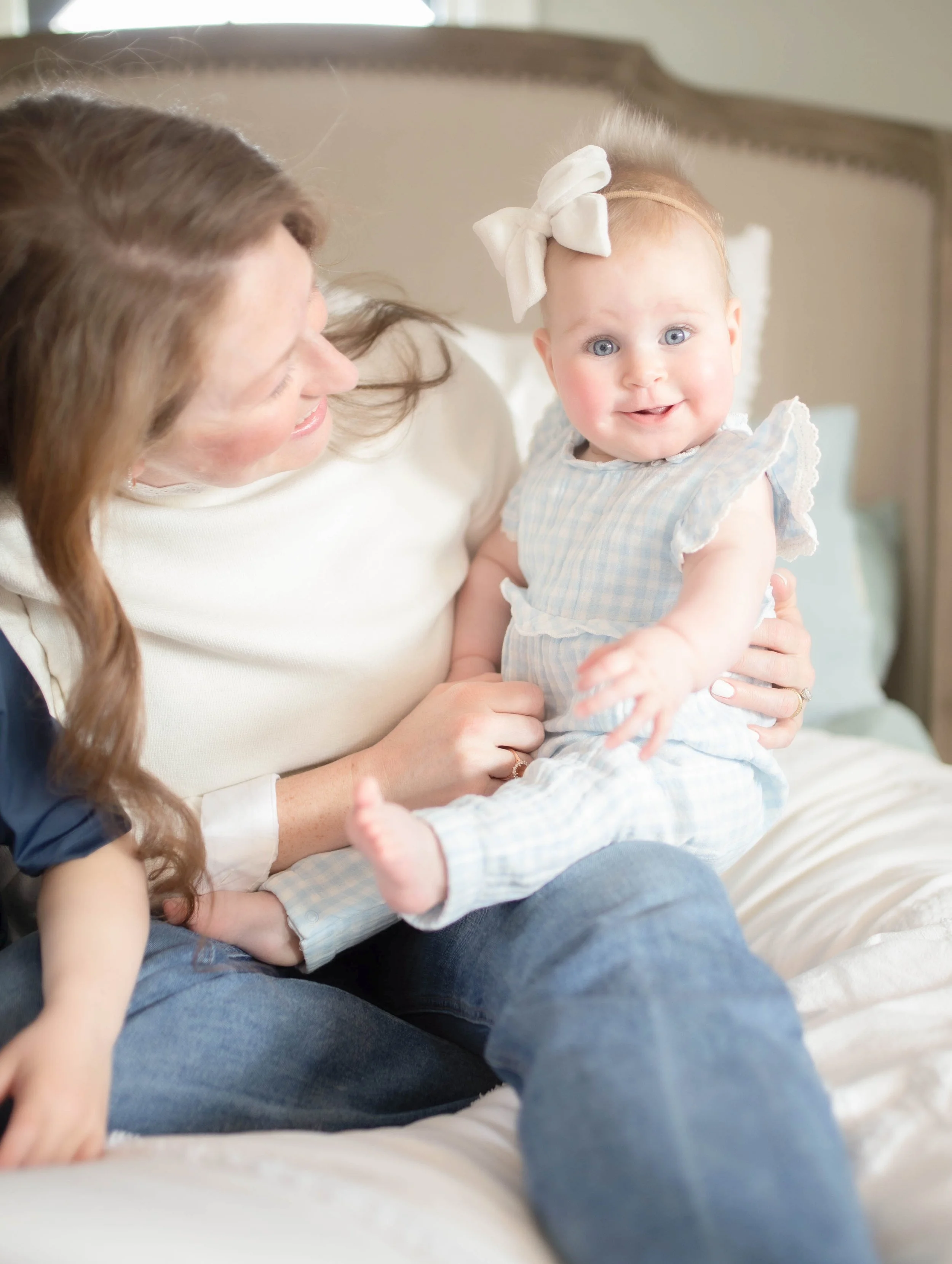 A woman with long brown hair holding a smiling baby girl with a white bow in her hair, sitting on a bed in a cozy bedroom.