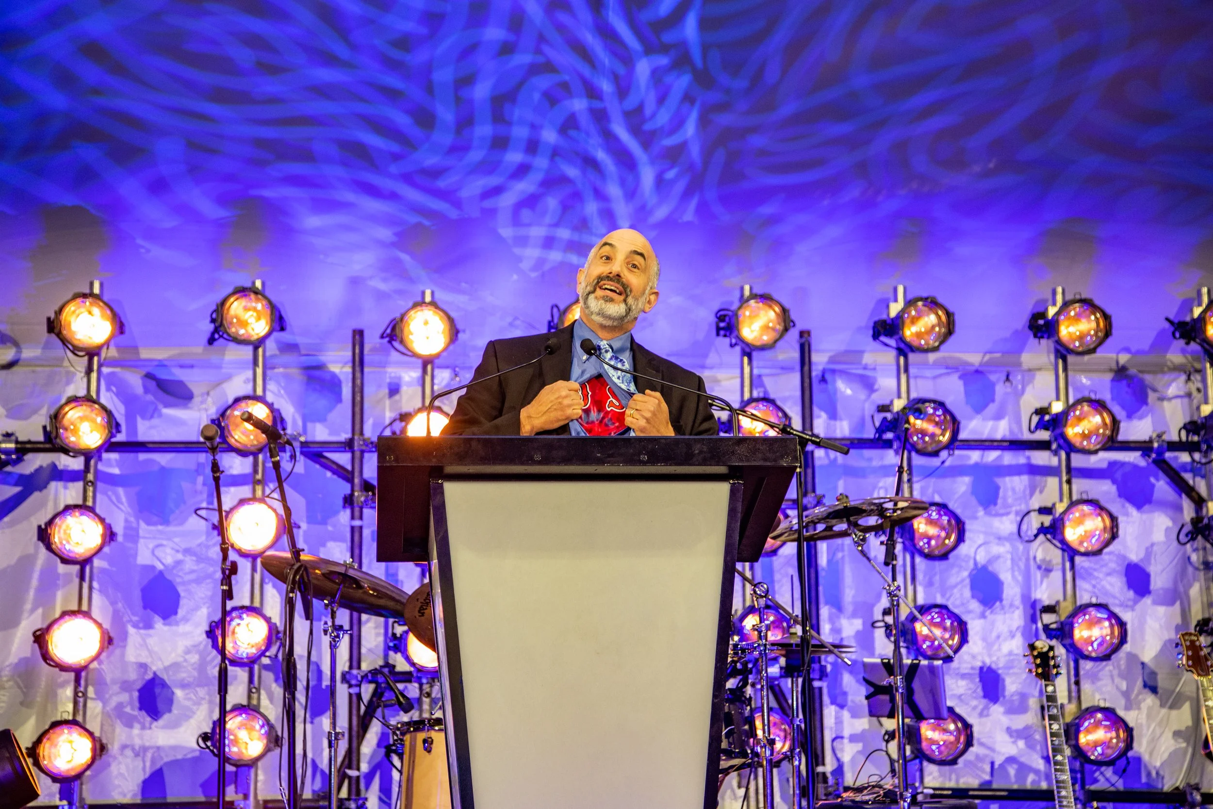 A man with a beard and gray hair stands at a podium, speaking at an event, with stage lights and guitars in the background. The stage has blue lighting and a decorated backdrop.