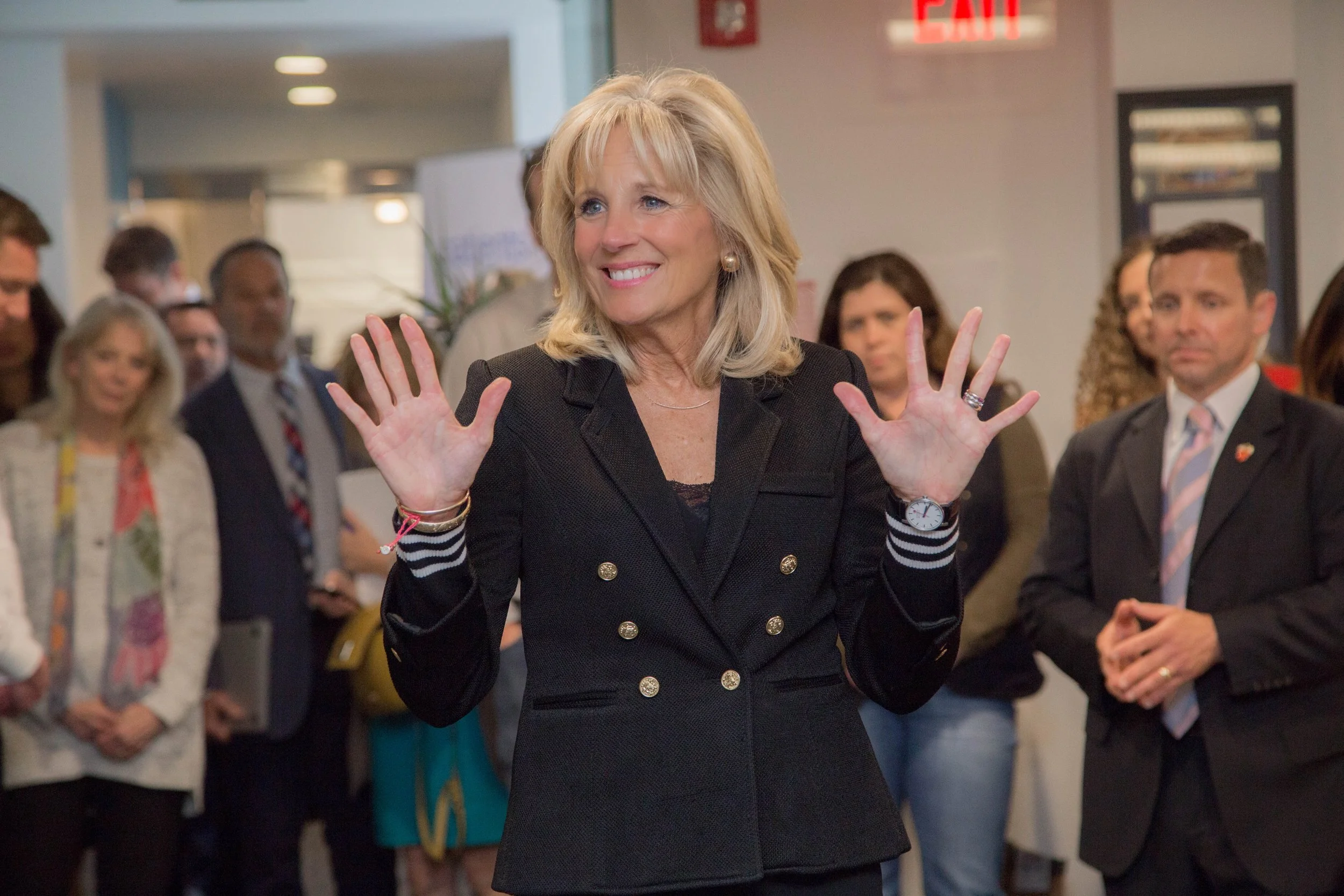 A smiling blonde woman in a black blazer with gold buttons gestures with her hands while surrounded by people at an indoor event.