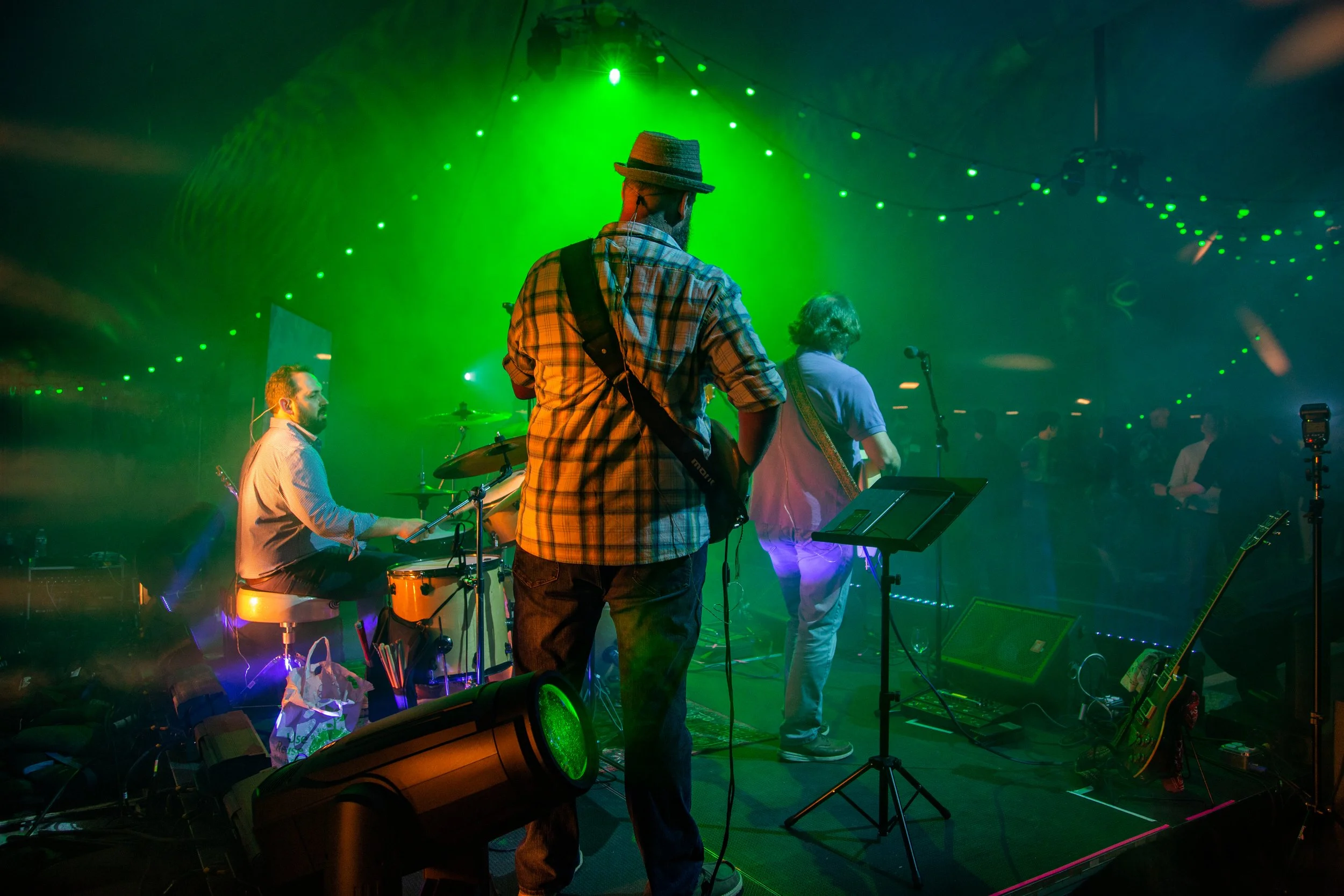 A band performing on stage with green lighting, including a drummer, a guitarist with a hat, and a lead singer with curly hair. Audience is visible in the background.