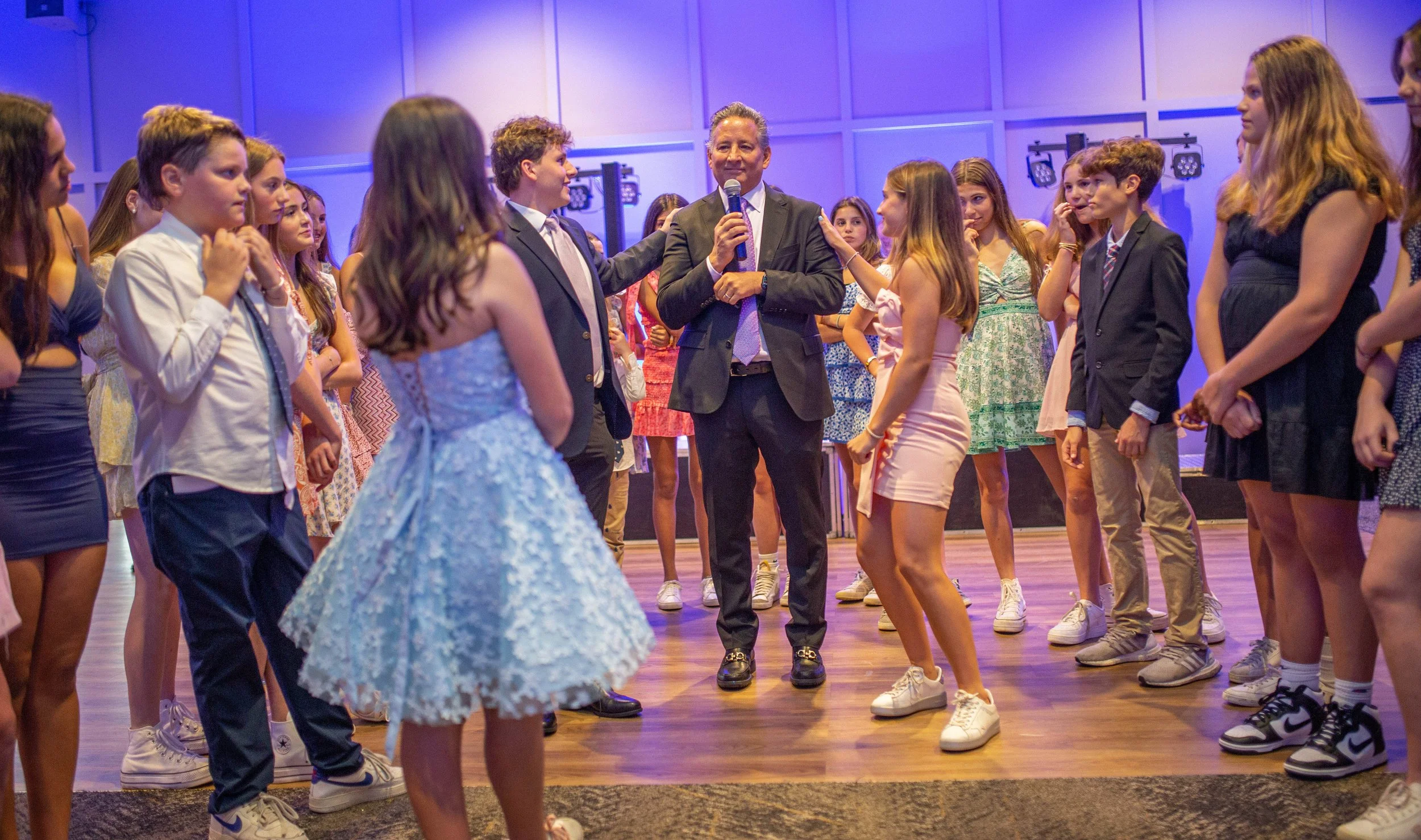 A group of children dancing in a circle on a wooden floor, with a man holding a microphone in the center, in a school or event setting with purple lighting and blue wall panels.