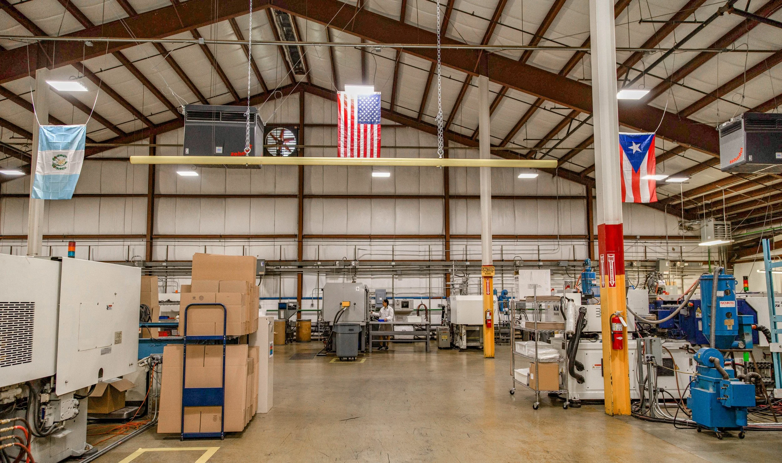 Interior view of a factory or warehouse with machinery, boxed goods, flags of the United States, Puerto Rico, and Guatemala hanging from the ceiling.