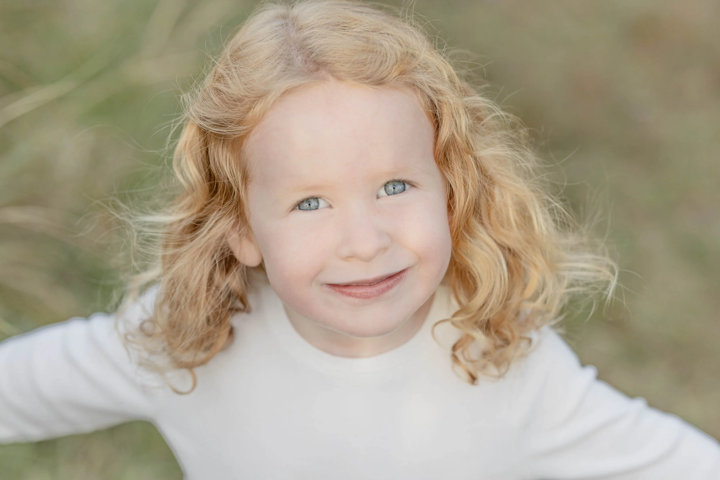 A young girl with curly red hair and blue eyes smiling outdoors.