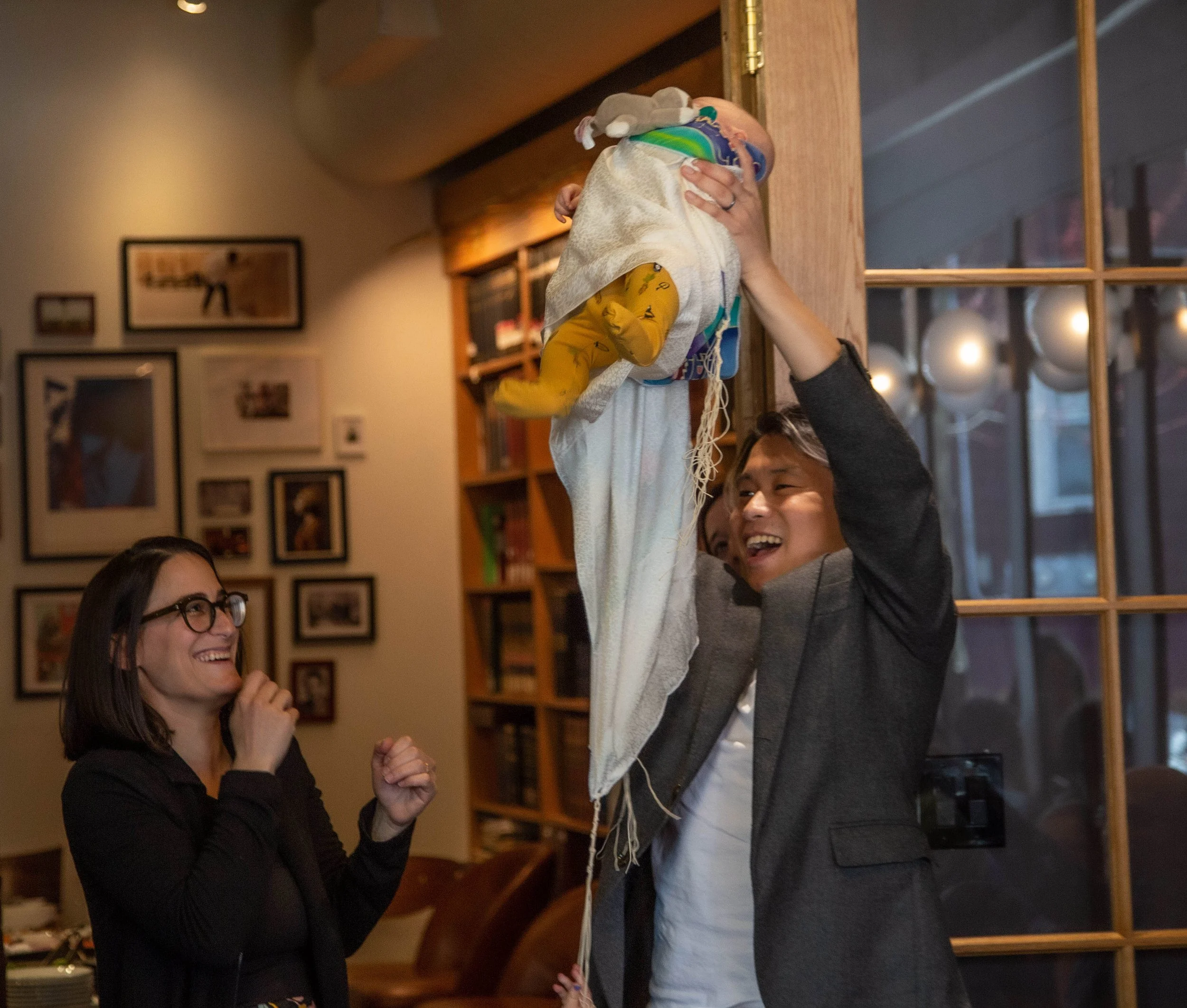 A man lifting a baby in a restaurant or cafe, smiling, with a woman observing and smiling in the background.