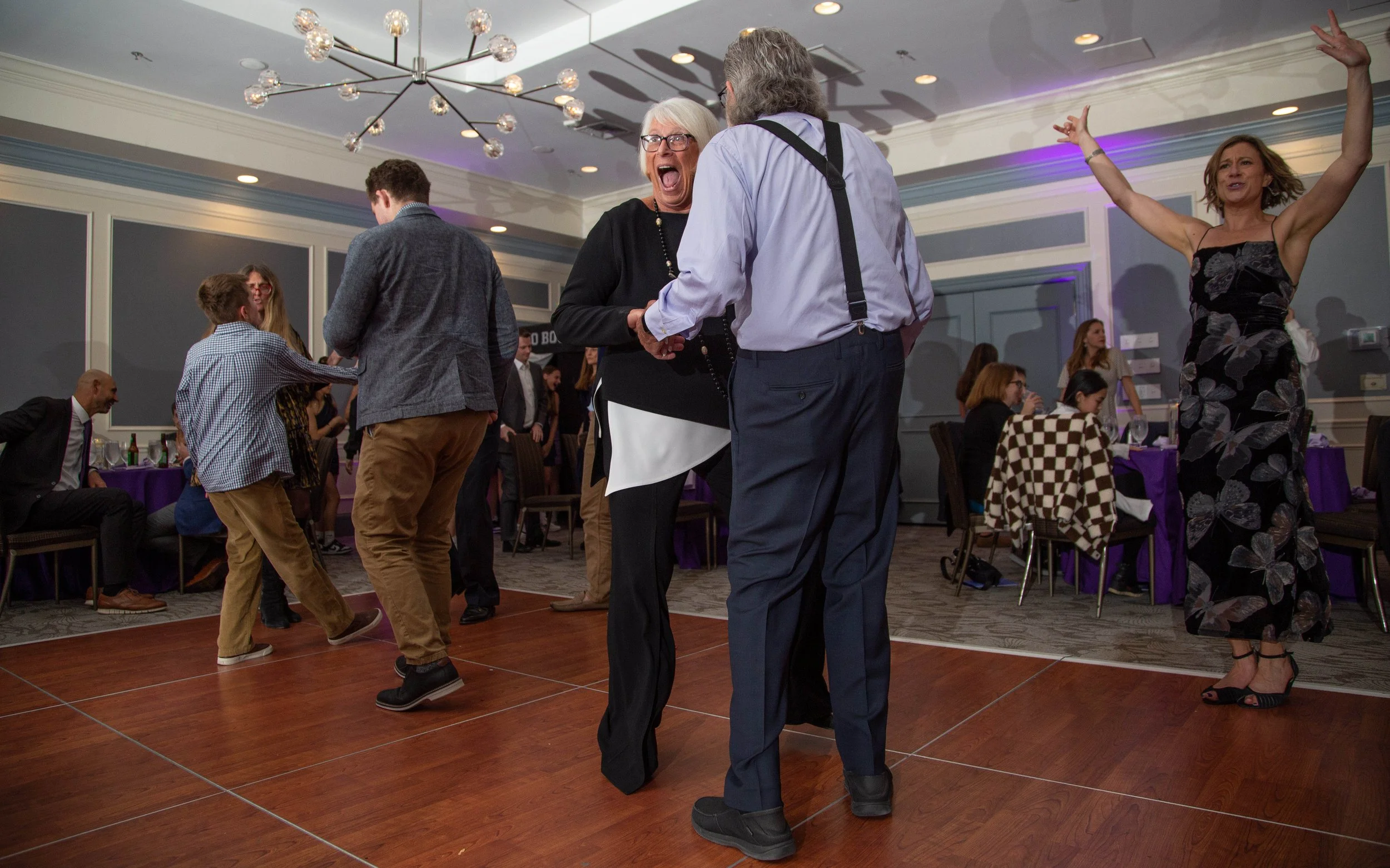 People dancing and celebrating at a party or wedding reception in a decorated ballroom with purple accents and modern lighting.
