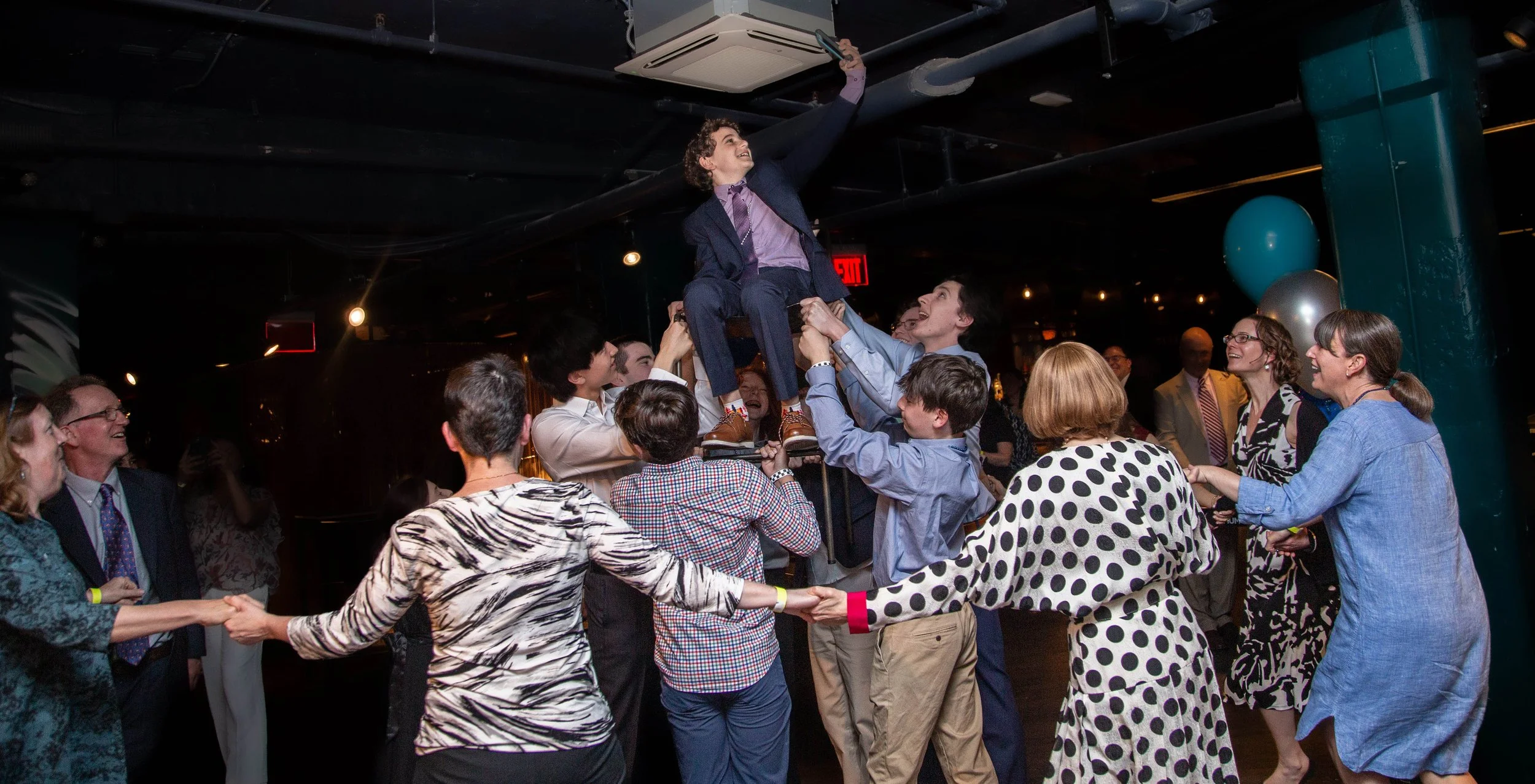 Group of people dancing and celebrating indoors, lifting and supporting a young boy in a suit who is sitting on a chair, surrounded by cheerful adults.