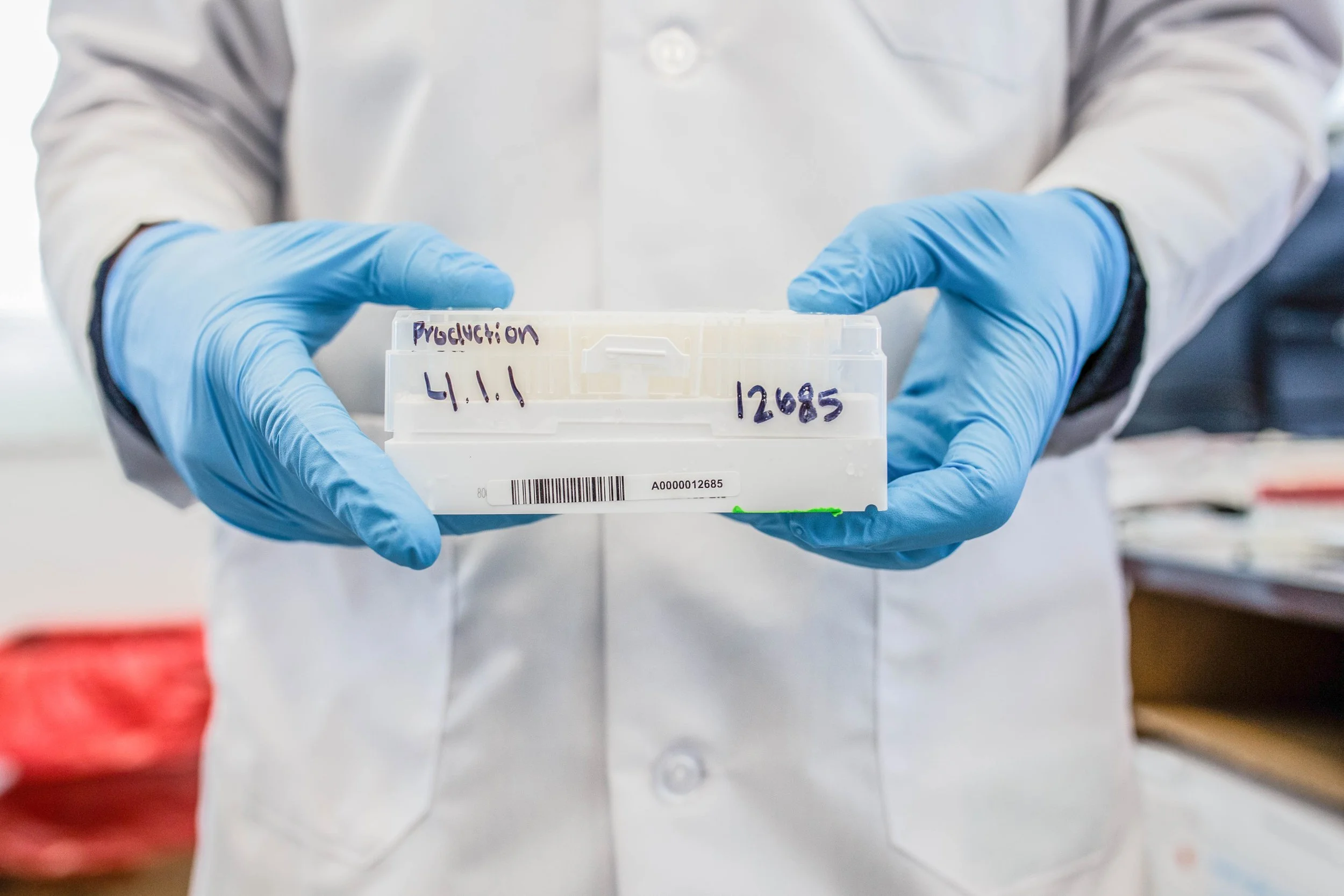 Person in a lab coat and blue gloves holding a clear plastic container with handwritten labels.