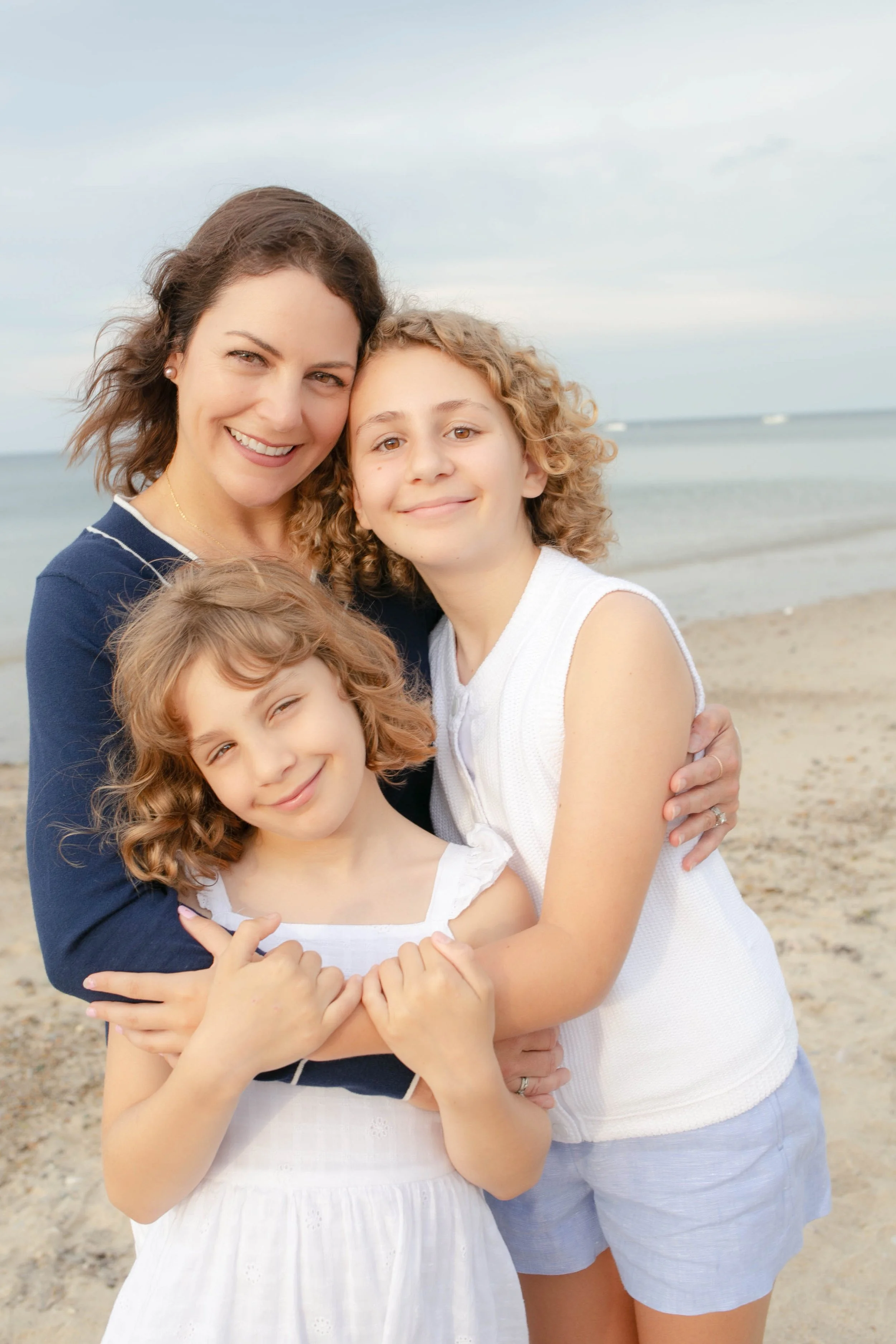 A woman and two young girls hugging on a beach with the ocean in the background.