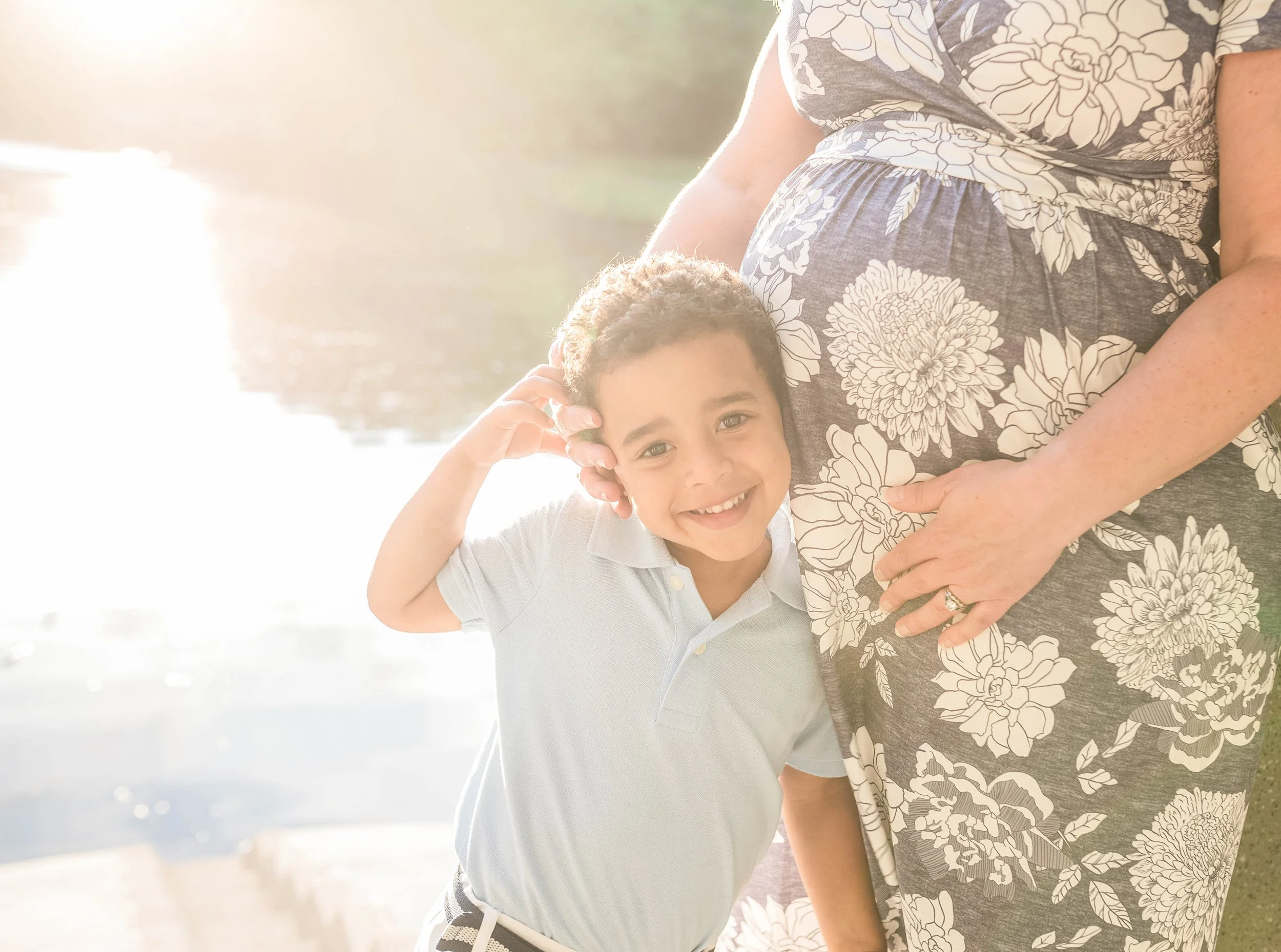 A smiling young boy standing next to a pregnant woman at a sunny outdoor location near water, with the boy touching the woman's pregnant belly.