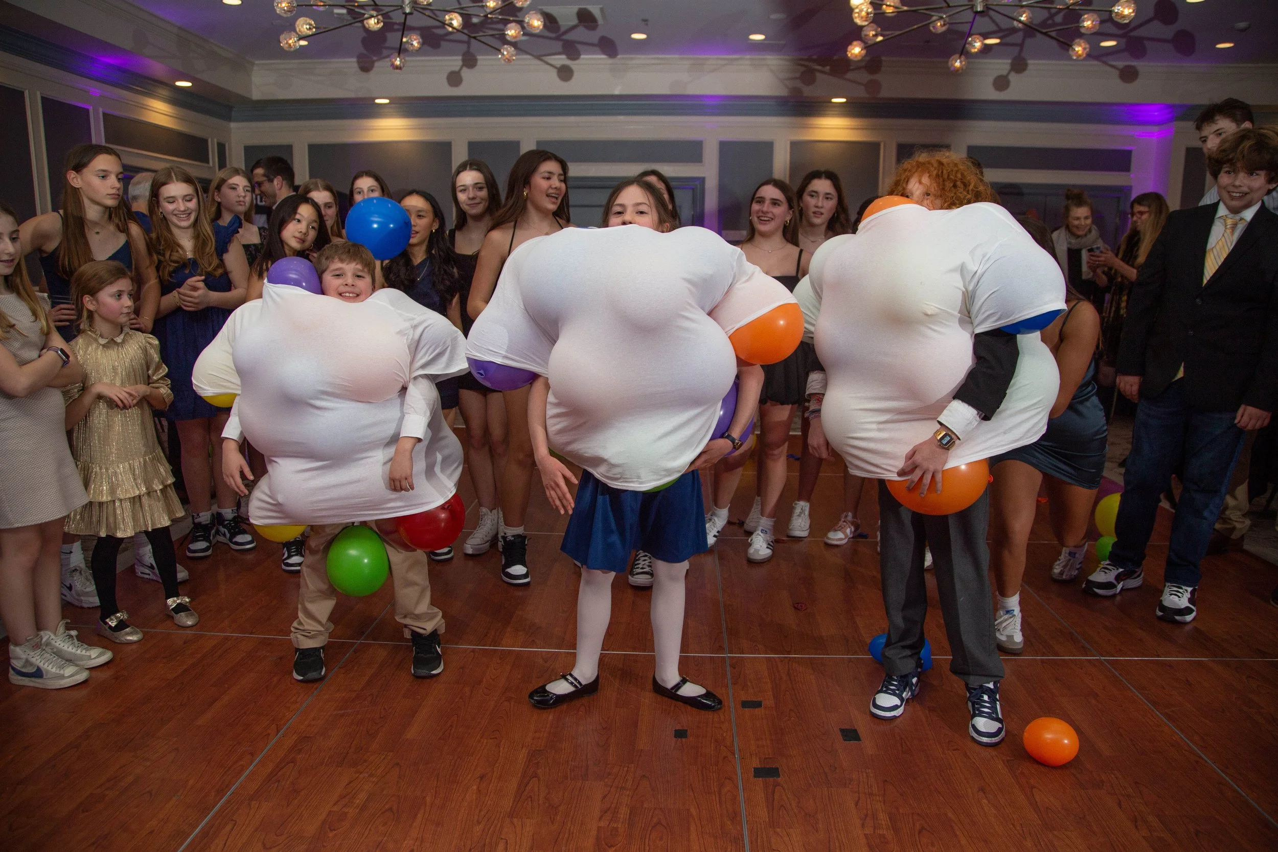 Children at a party wearing large costume shirts with balloons attached, playing a game on a wooden floor with adults and other kids watching and laughing in the background.