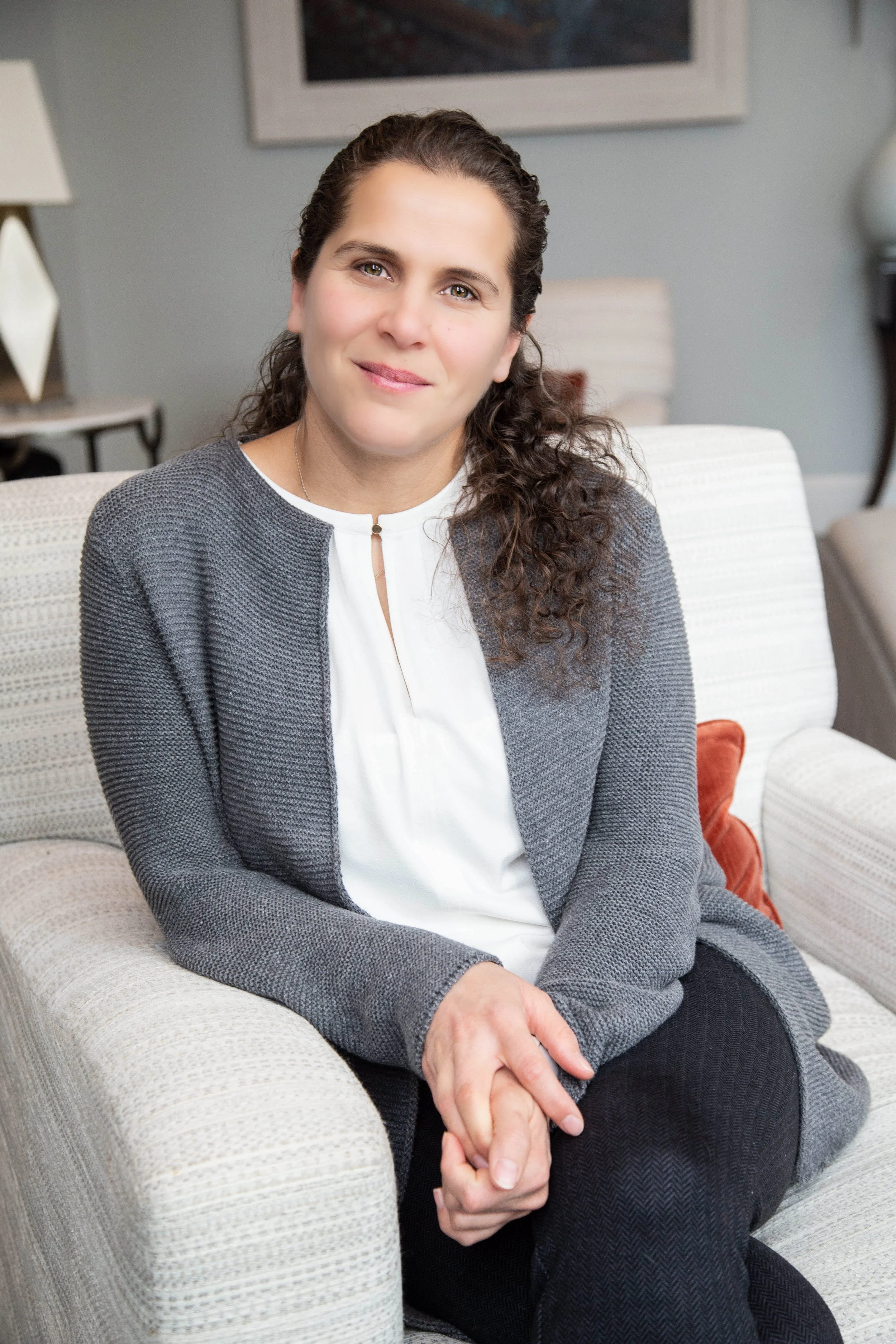 A woman with dark wavy hair sitting on a beige armchair in an indoor setting, wearing a gray jacket over a white top, looking at the camera with a slight smile.