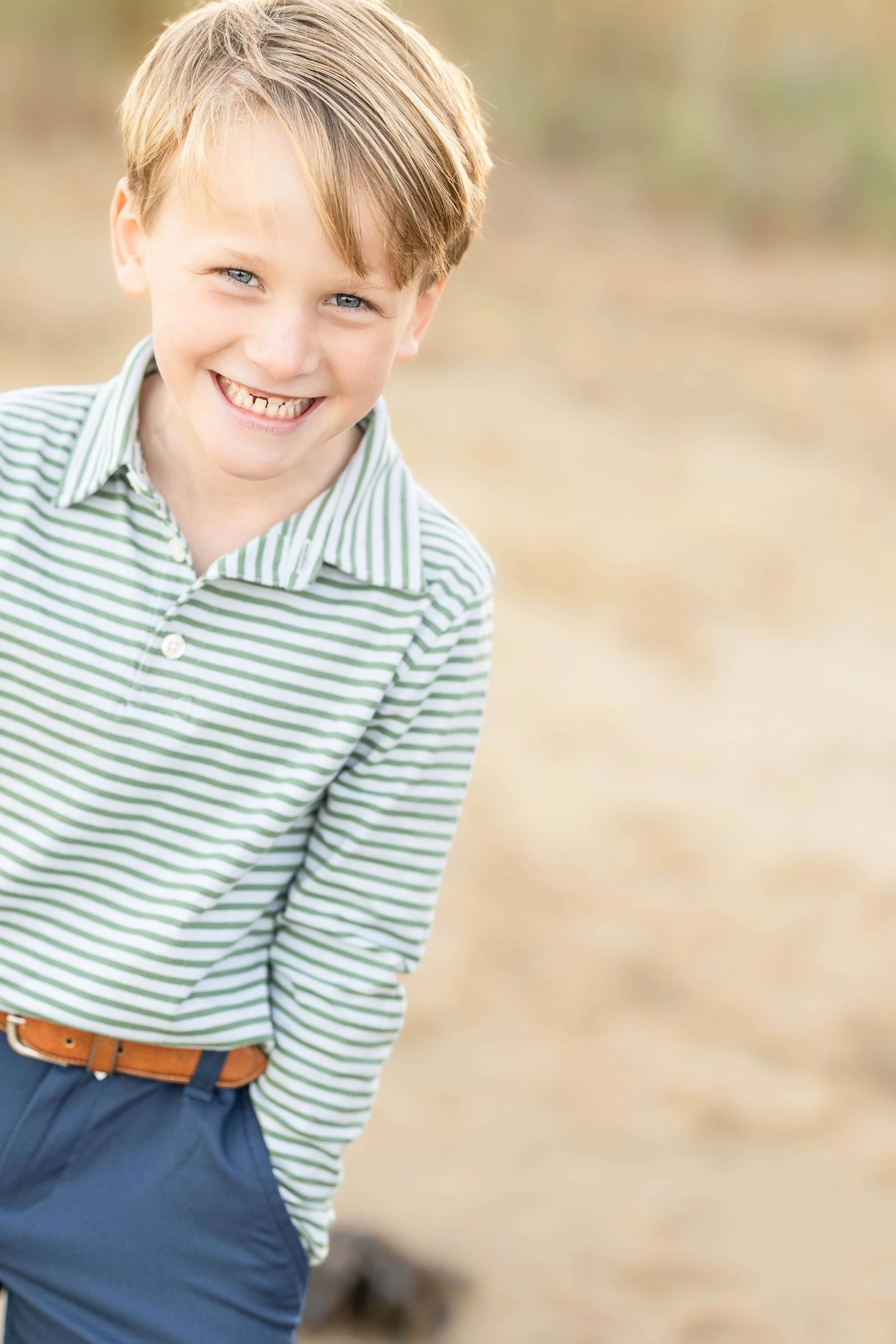 A young boy with blond hair and blue eyes smiling outdoors, wearing a green and white striped shirt and dark blue pants.