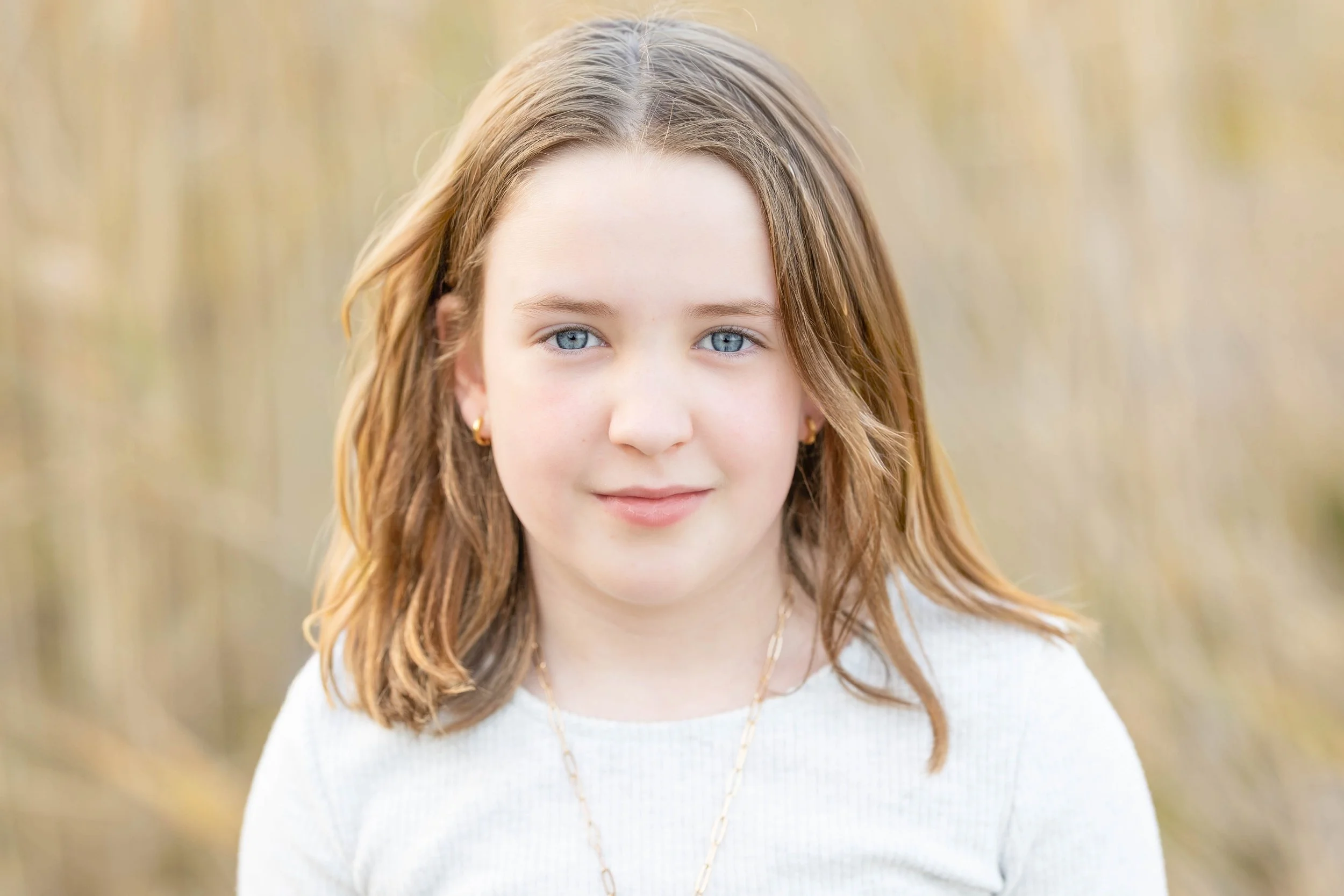 A young girl with light brown hair, blue eyes, and gold earrings is looking at the camera in a natural outdoor setting.