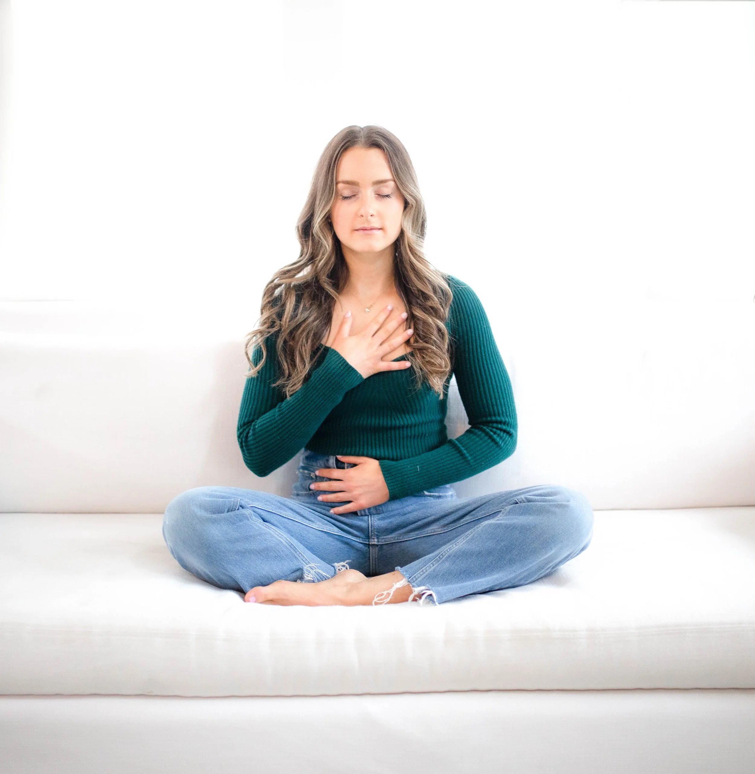 A woman sitting cross-legged on a white sofa with her eyes closed, one hand on her chest, and the other on her stomach, appearing to be meditating or practicing mindfulness.