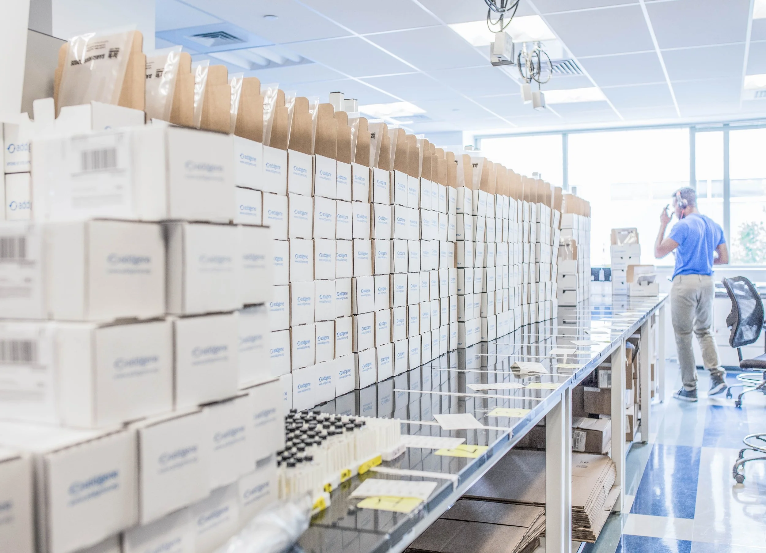 A man in a blue shirt and beige pants talking on the phone inside a room filled with stacks of boxes, some on a table and some on shelves, with large windows in the background letting in natural light.