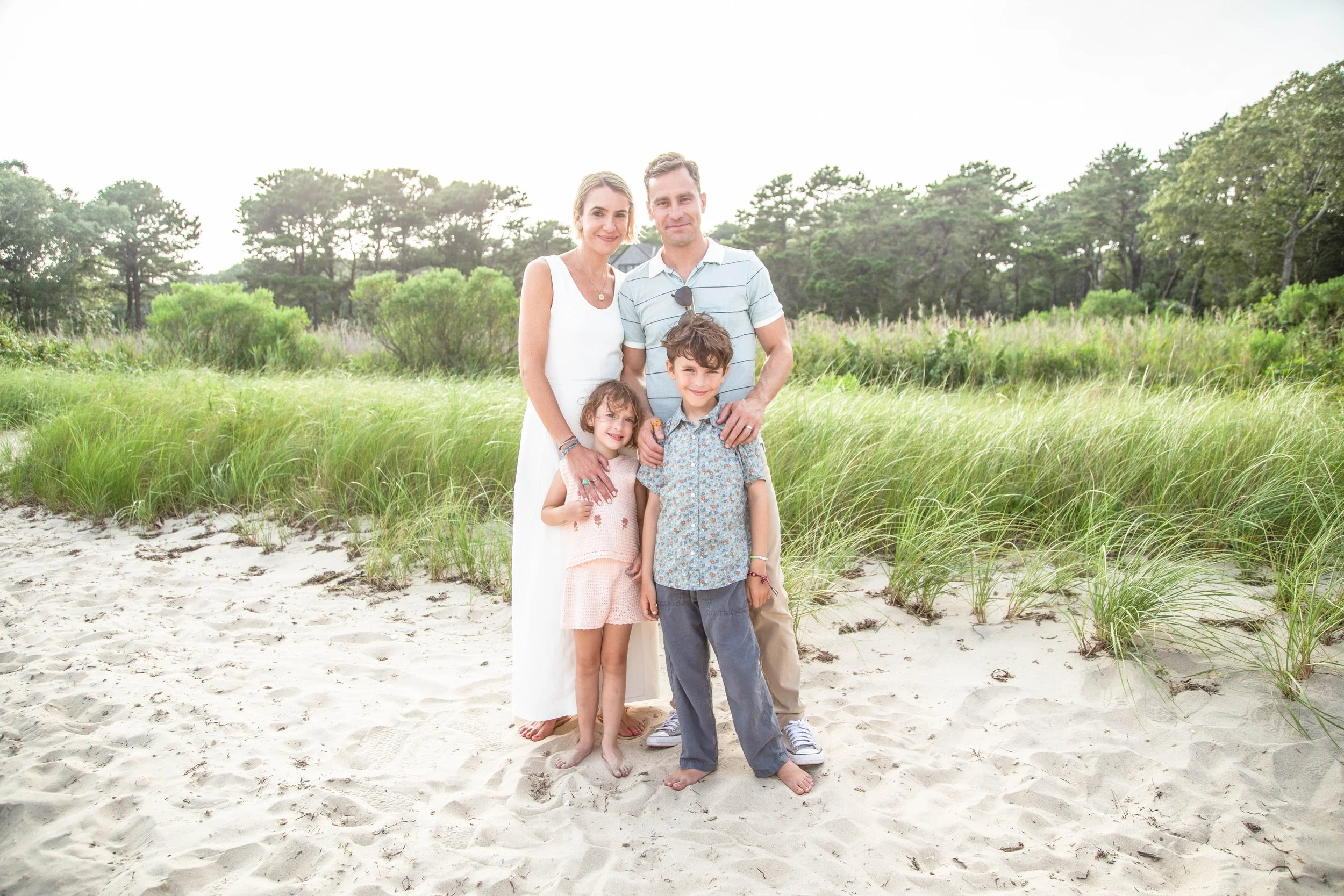 A family of four standing barefoot on a sandy beach with green grass and trees in the background, smiling towards the camera.