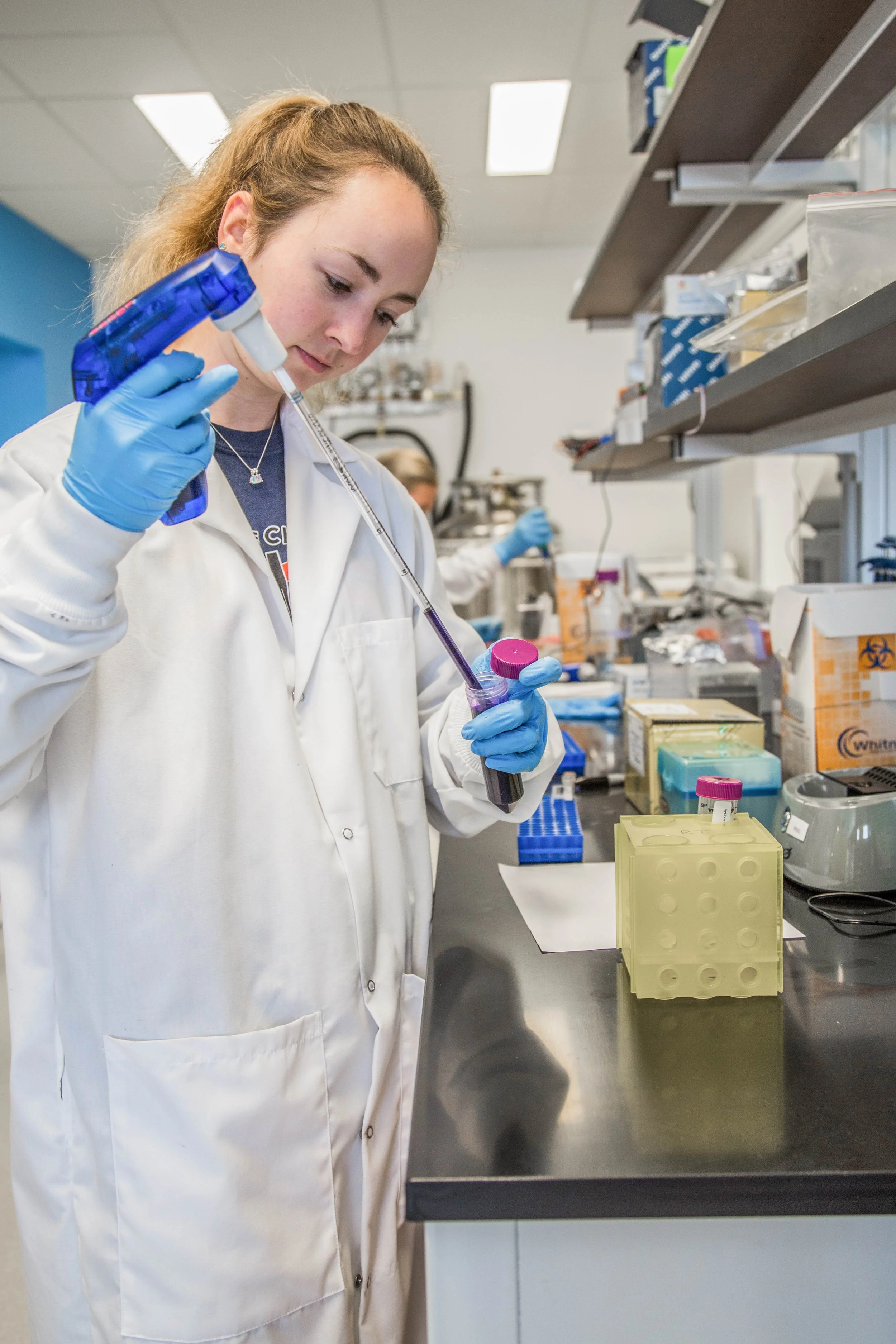 A female scientist in a white lab coat and blue gloves is pipetting a liquid into a test tube in a laboratory.
