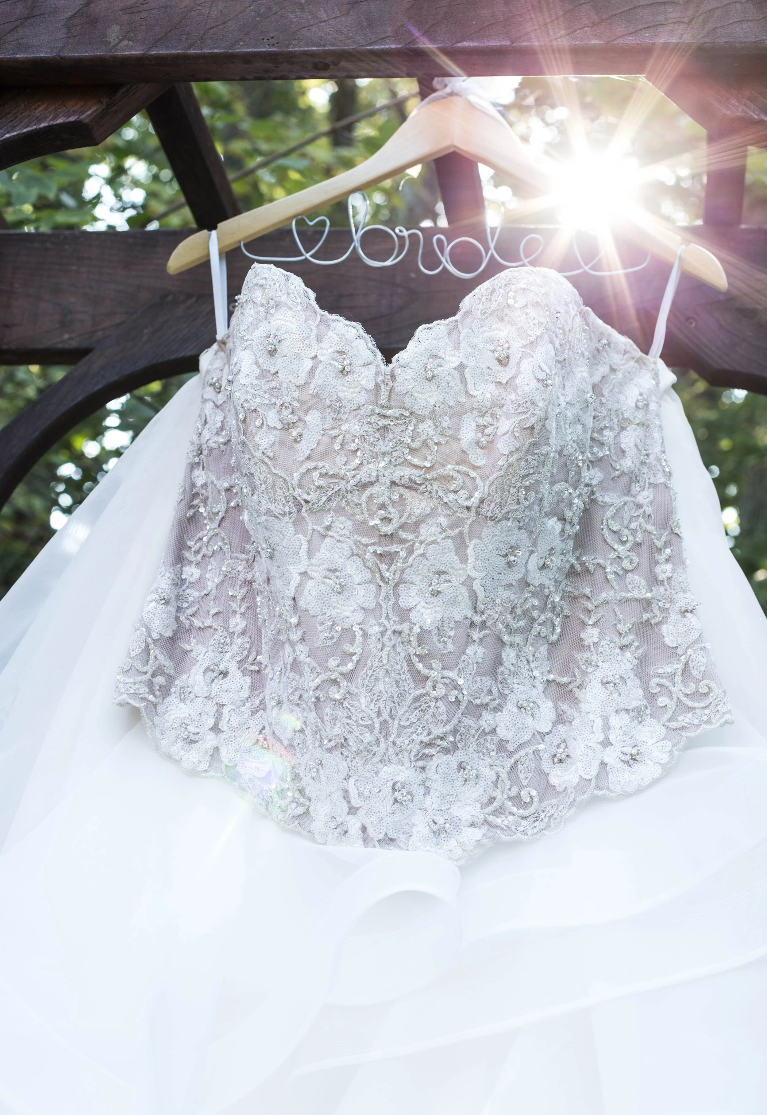 A white wedding dress with intricate lace and beaded embroidery hanging on a wooden hanger, with sunlight shining through, outdoors on a sunny day.