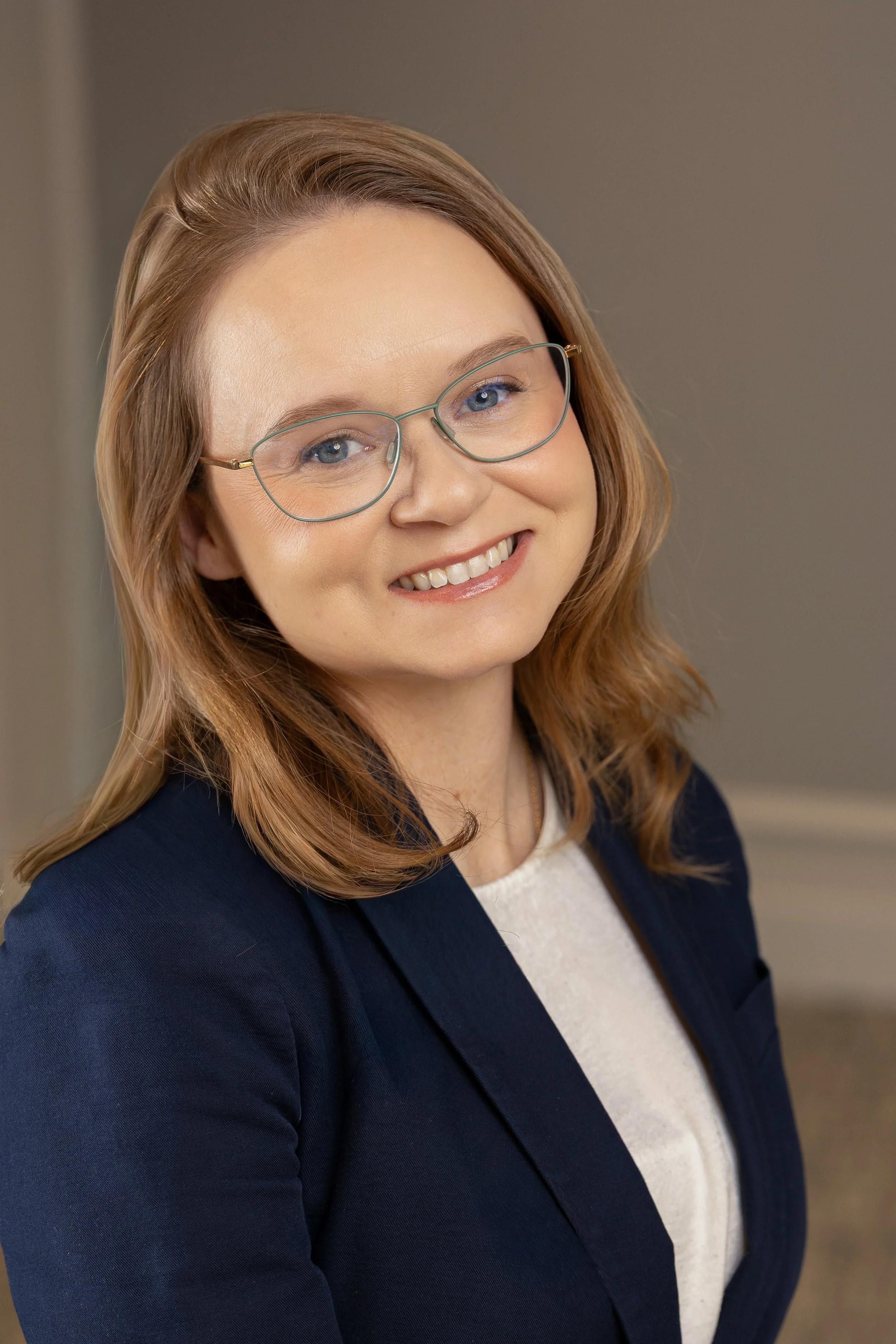 A smiling woman with shoulder-length reddish-brown hair, wearing glasses, a navy blazer, and a white top, standing indoors with a plain background.