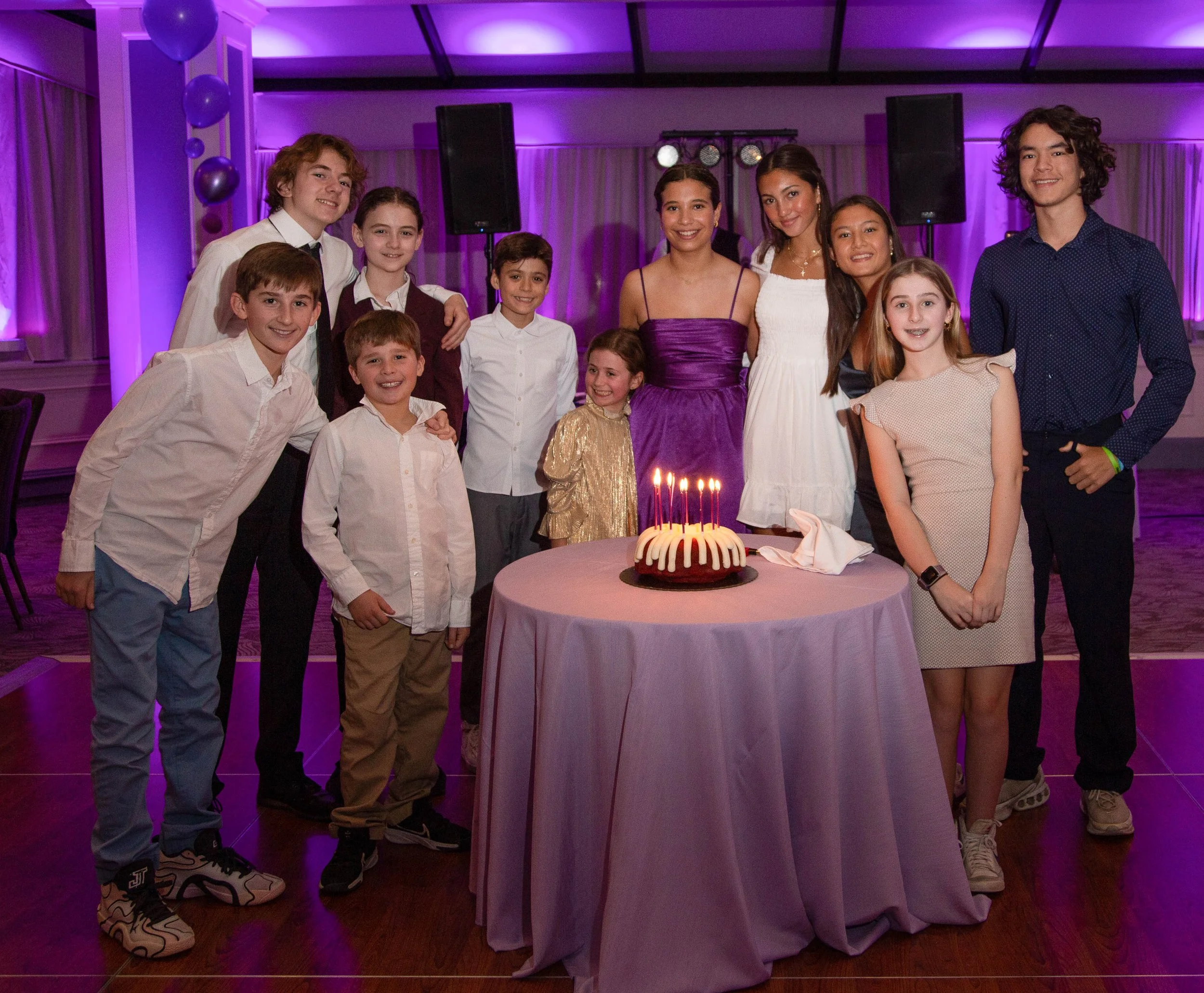 A group of children and teenagers gathered around a birthday cake with lit candles, at a celebration in a decorated hall with purple lighting.