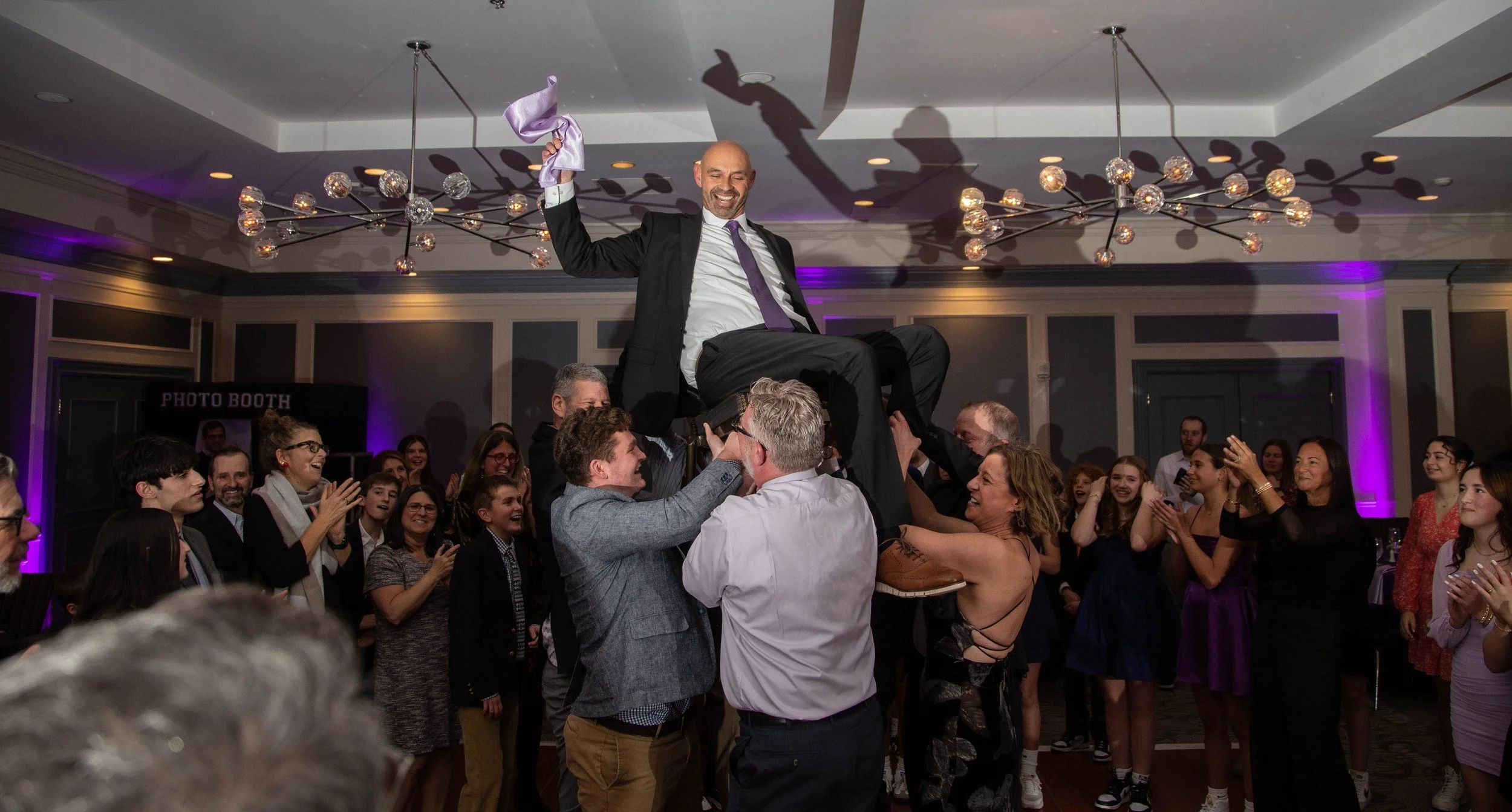 Groom being lifted on a chair during a wedding reception celebration surrounded by guests clapping and smiling.