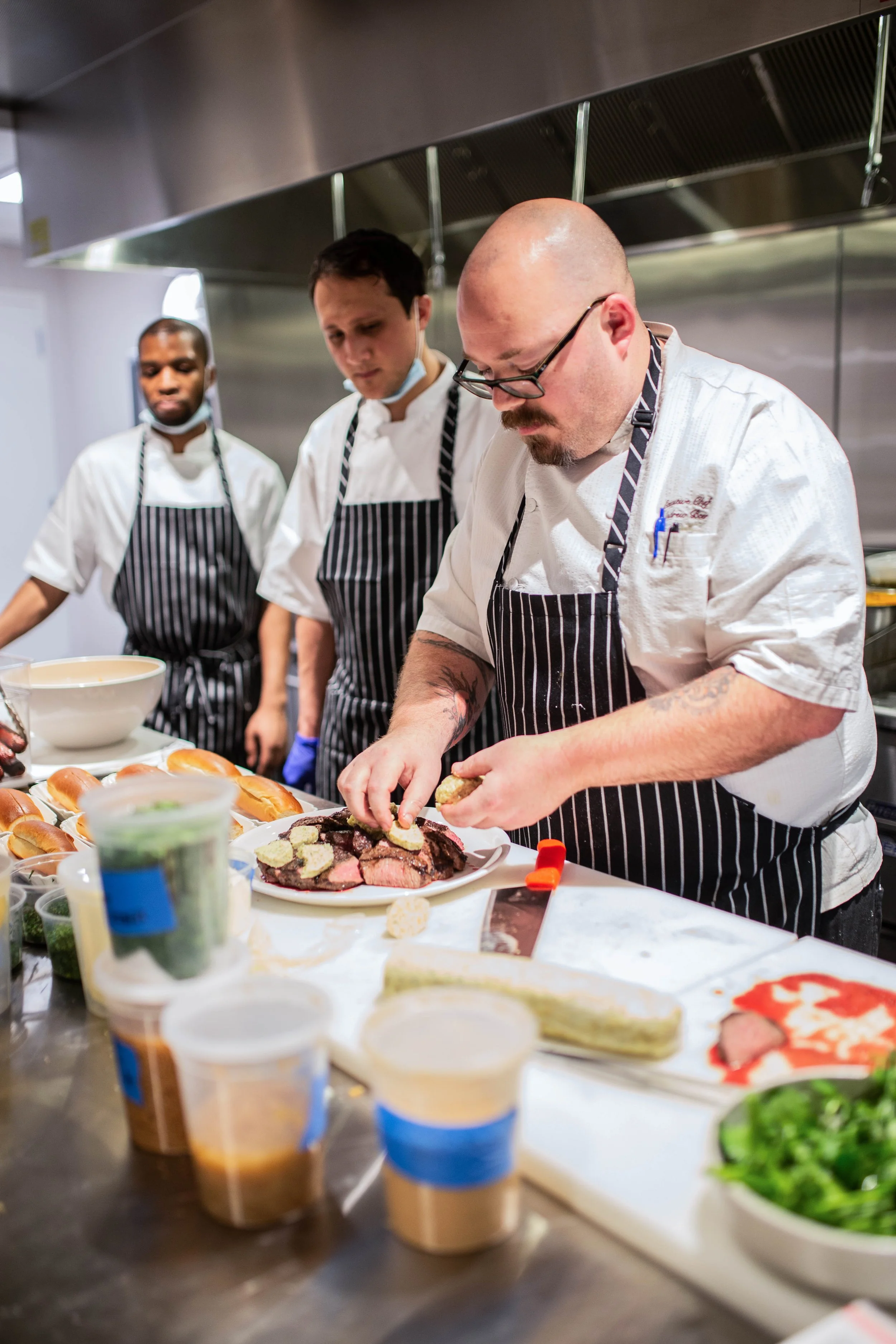 Three chefs in striped aprons preparing dishes in a professional kitchen, with one garnishing a plate of sliced steak.