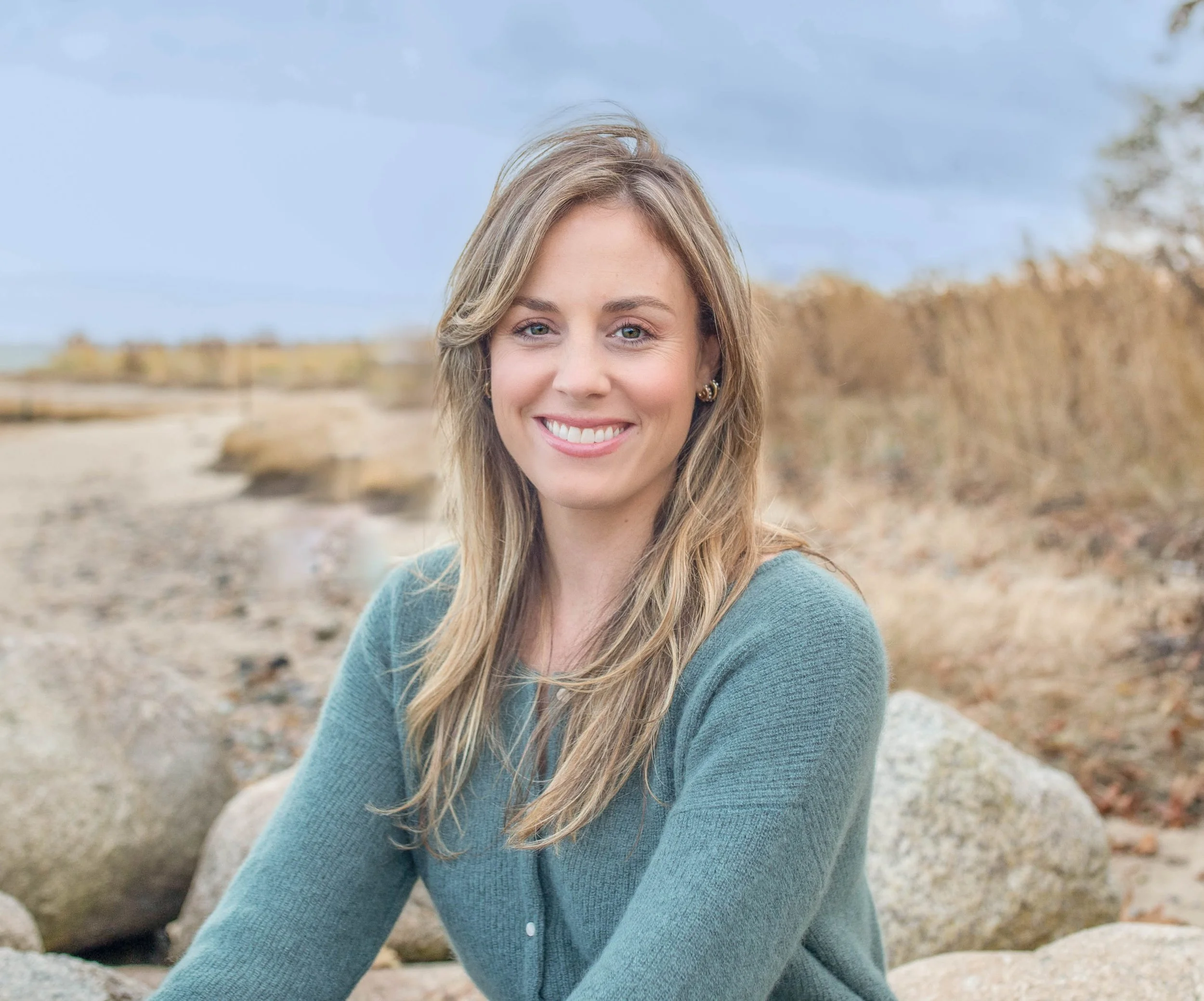 A smiling woman with long light brown hair, wearing a teal sweater, sitting on rocks outdoors with a beach and trees in the background.