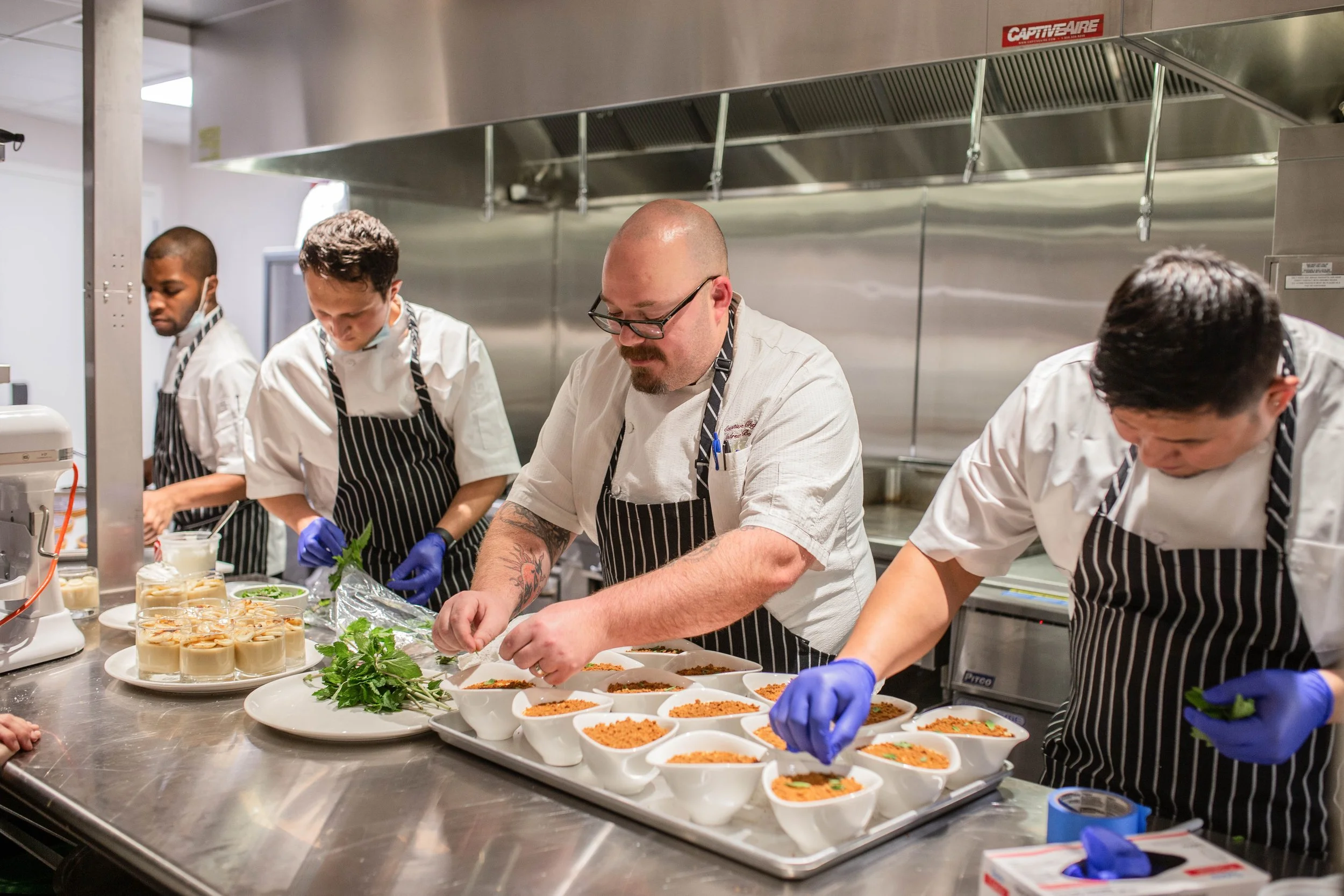 A group of five chefs in a professional kitchen preparing and garnishing dishes, with one chef in front and others in the background.