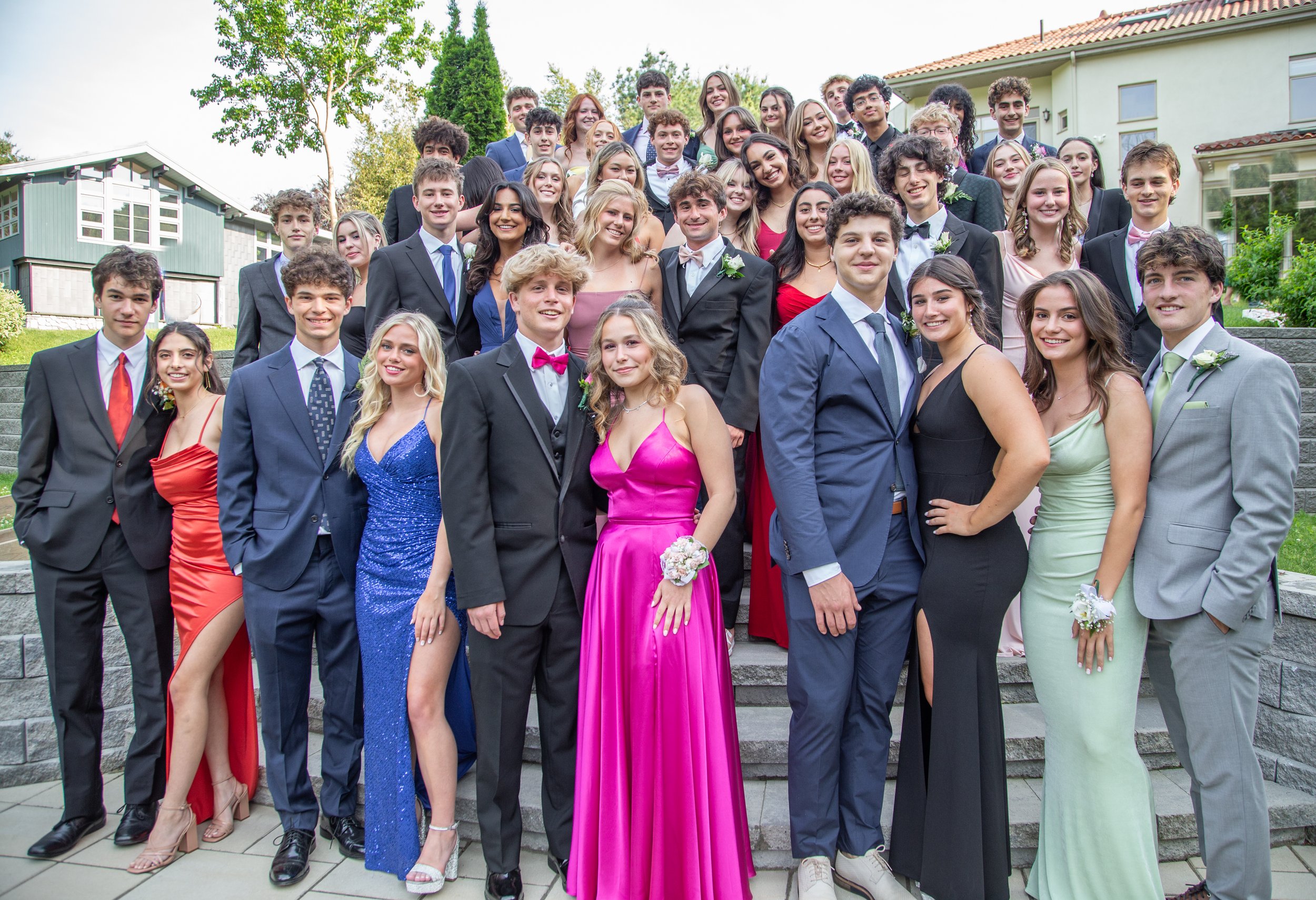 Group of young people dressed in prom attire outdoors, standing on steps with greenery and houses in background, smiling at the camera.