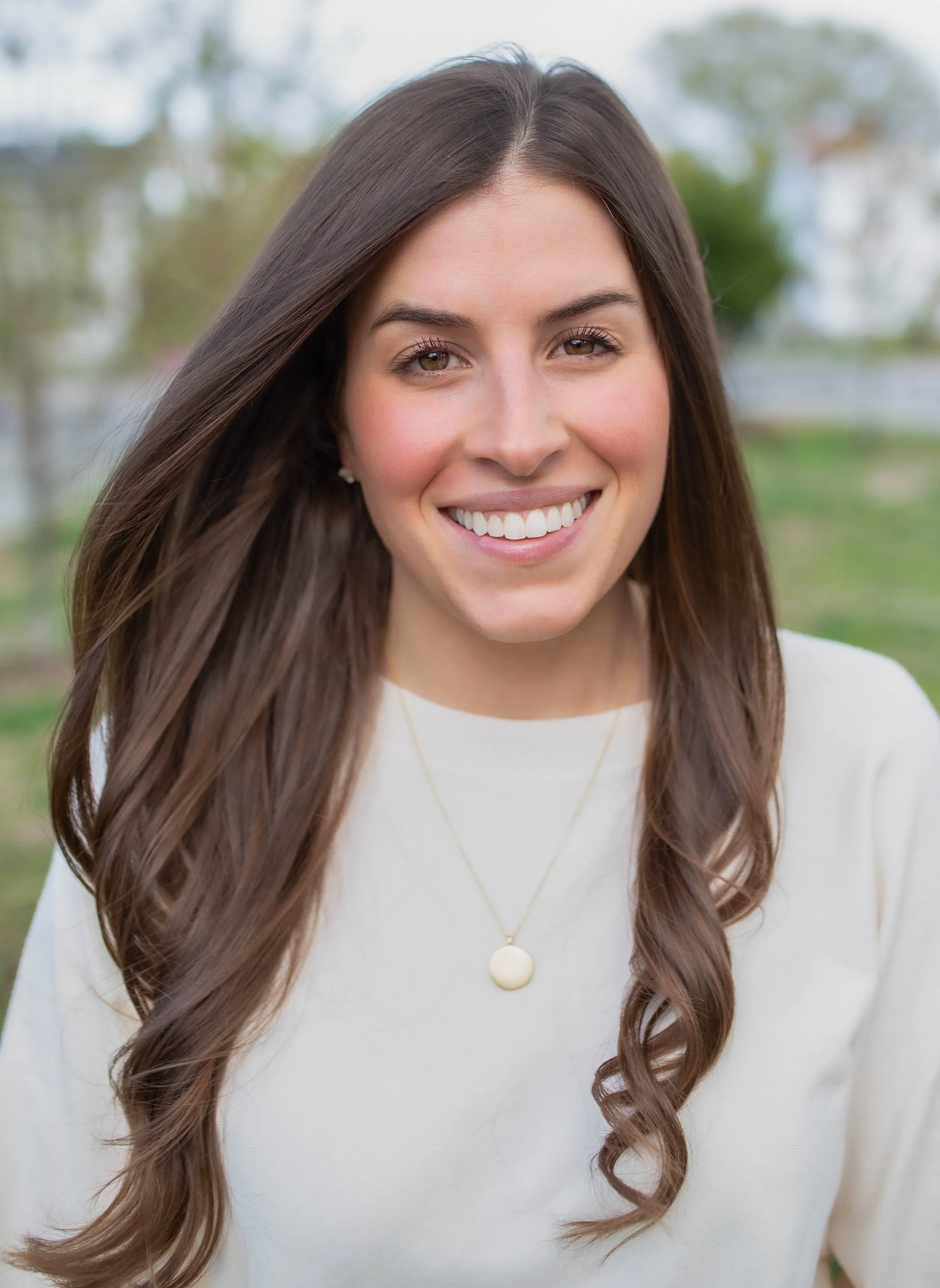 A young woman with long brown hair smiling outdoors, wearing a white top and a gold necklace with a circular pendant.