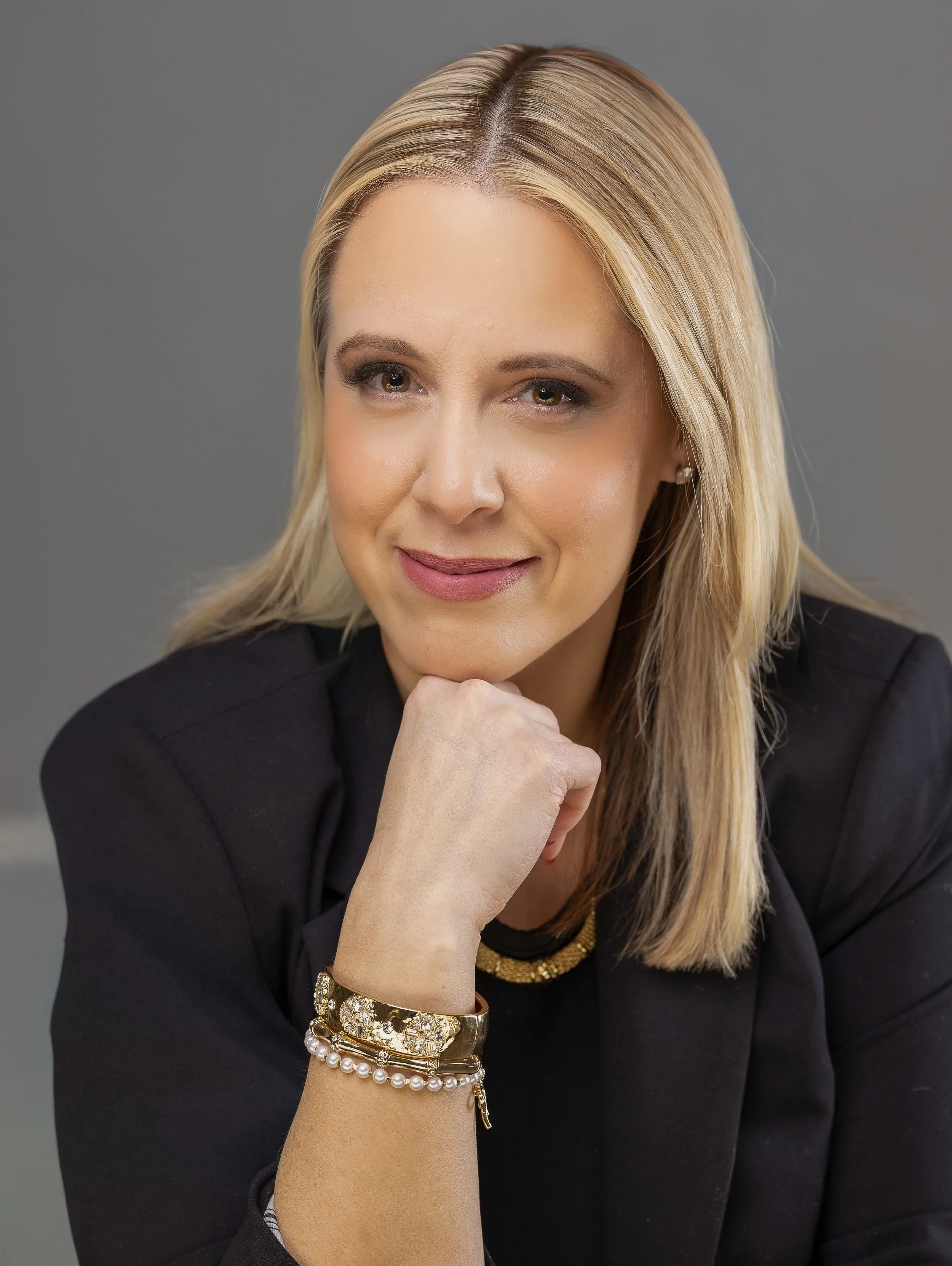 A woman with blonde hair in a black blazer, resting her chin on her hand, wearing gold and pearl jewelry, smiling softly against a gray background.