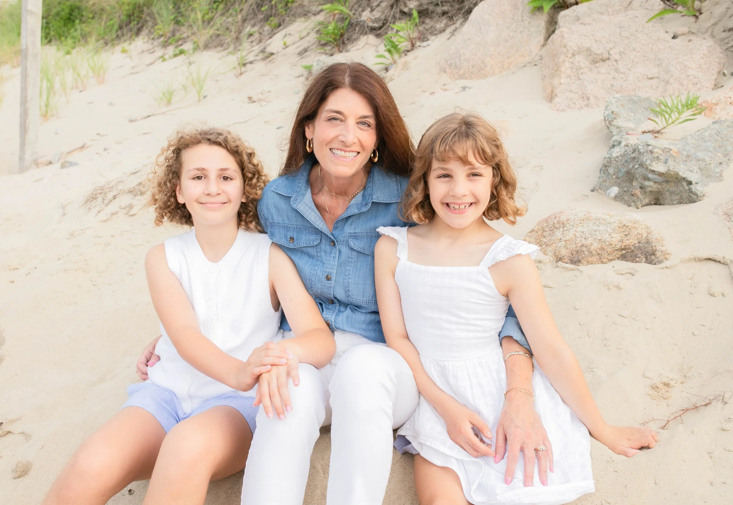 A woman with two young girls sitting on the sand at the beach, smiling at the camera.