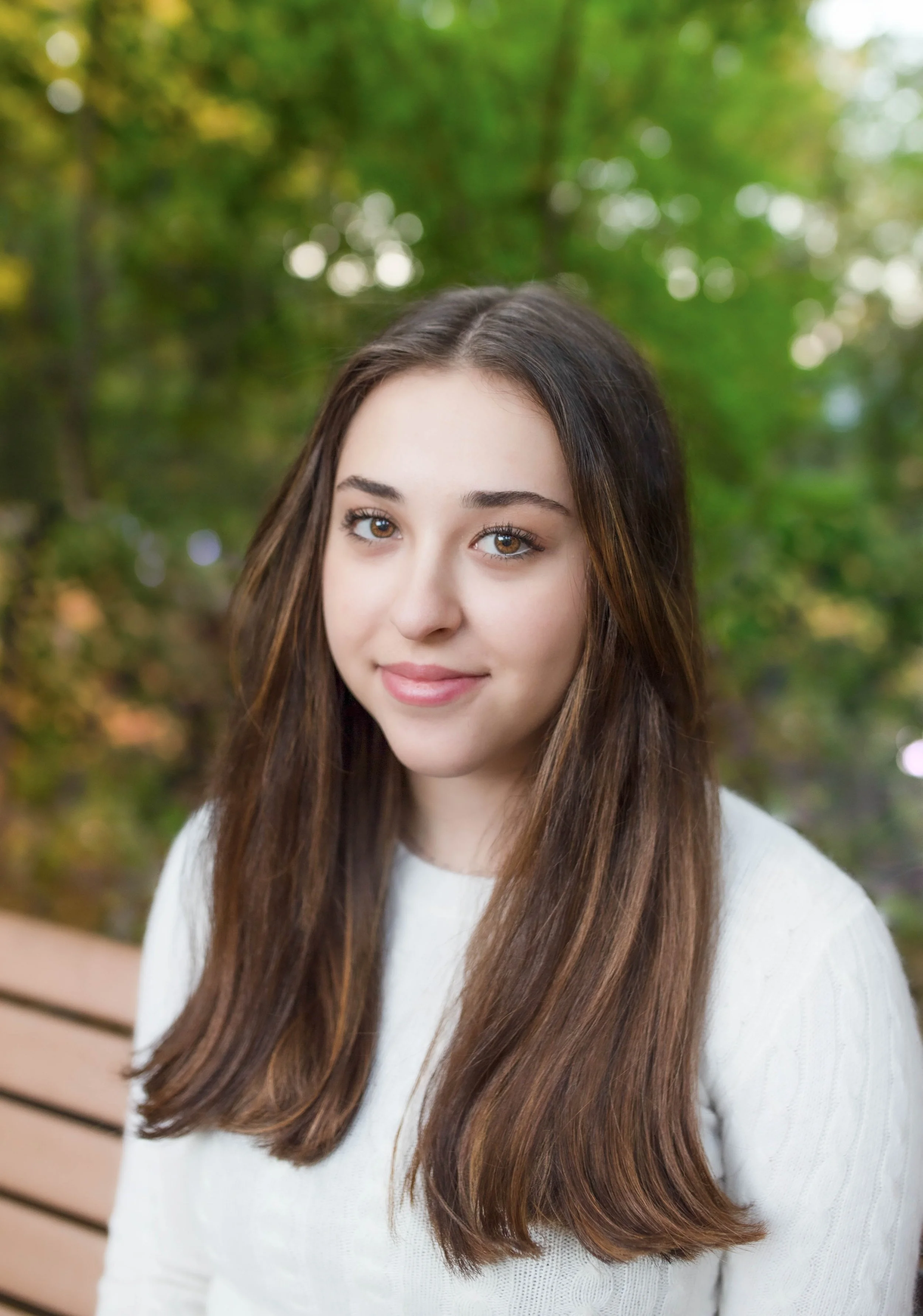 A young woman with long brown hair, wearing a white sweater, sitting on a park bench with green trees in the background.