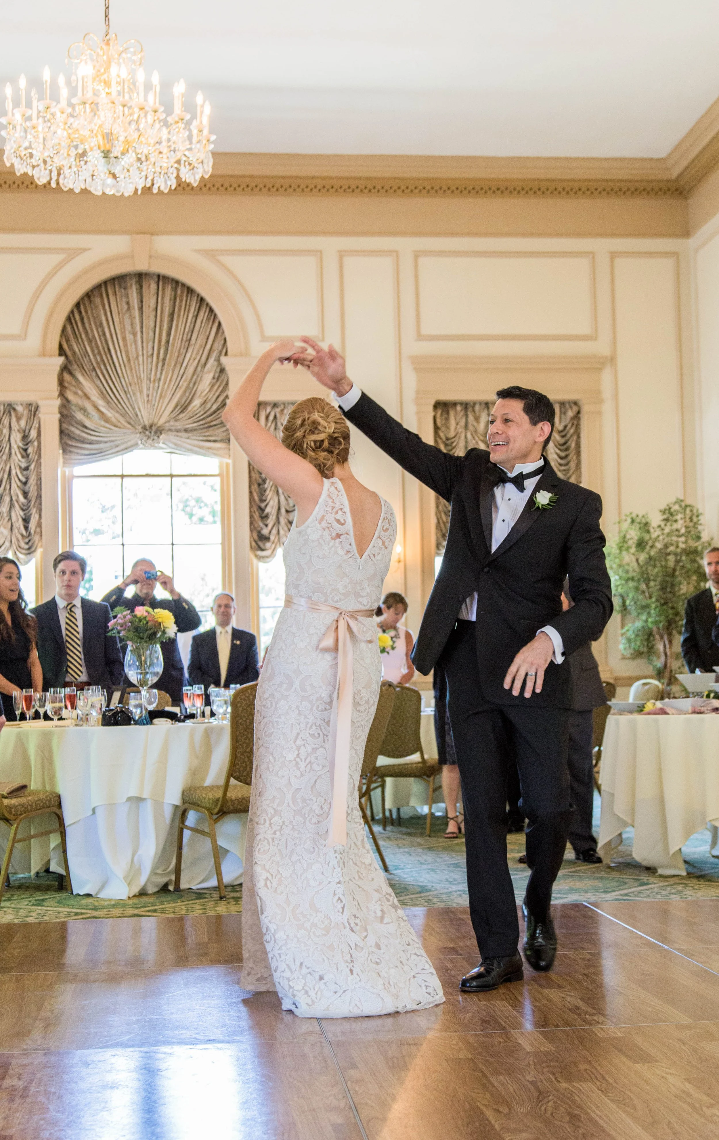A bride and groom dancing at a wedding reception in a ballroom with a chandelier and large windows.