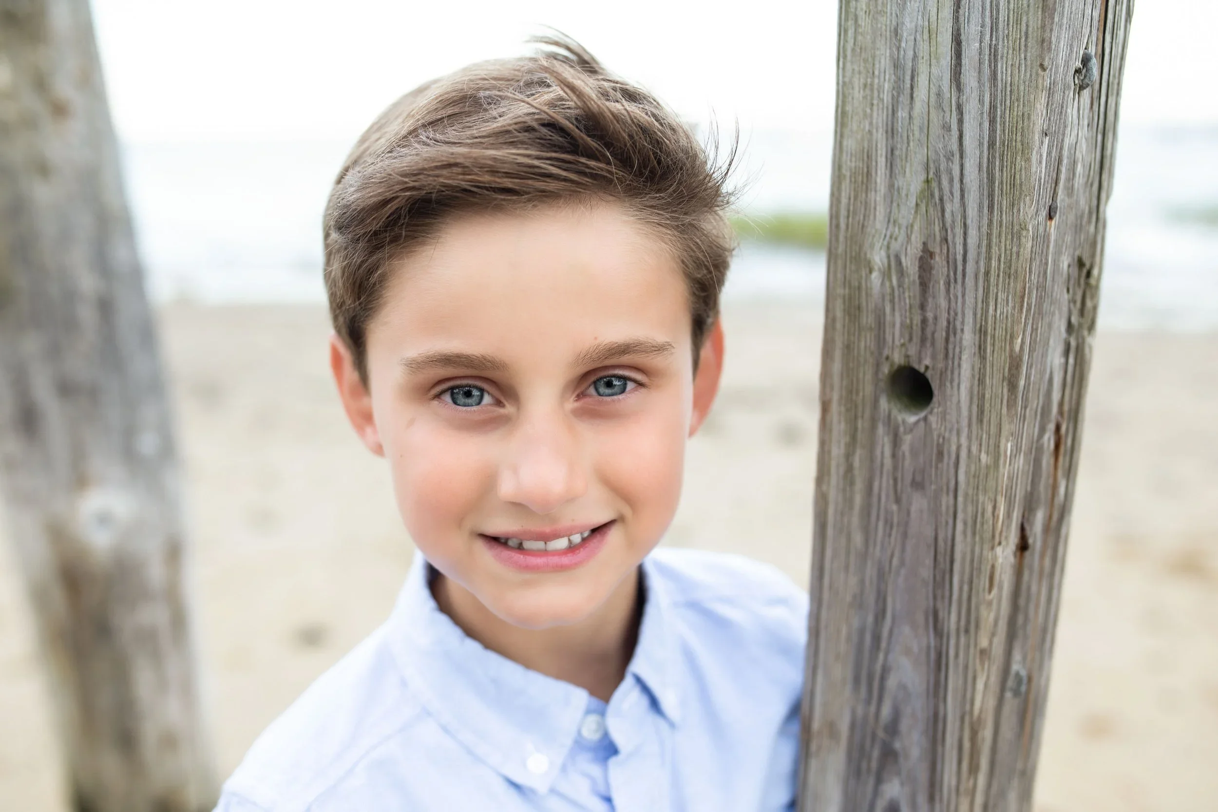 A young boy with blue eyes and light brown hair peeking through two wooden posts at a beach.