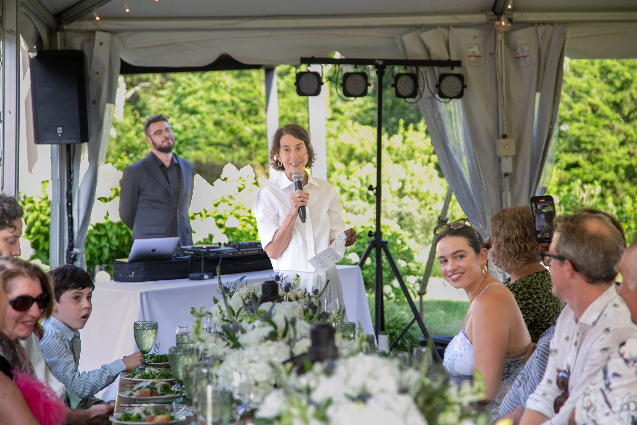 A woman speaking into a microphone at an outdoor gathering under a tent, with a DJ and green foliage in the background, and guests seated at a decorated table with white flowers.