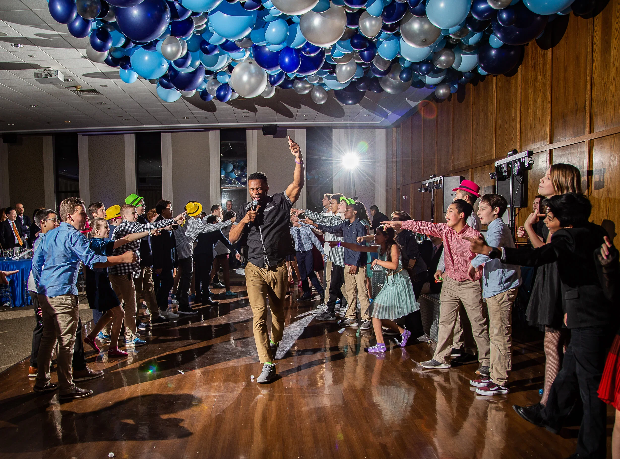 A lively dance party with kids and a DJ in a banquet hall decorated with blue and silver balloons hanging from the ceiling. A man in the center is dancing with a microphone, leading the group with energetic moves. The children are dressed in colorful