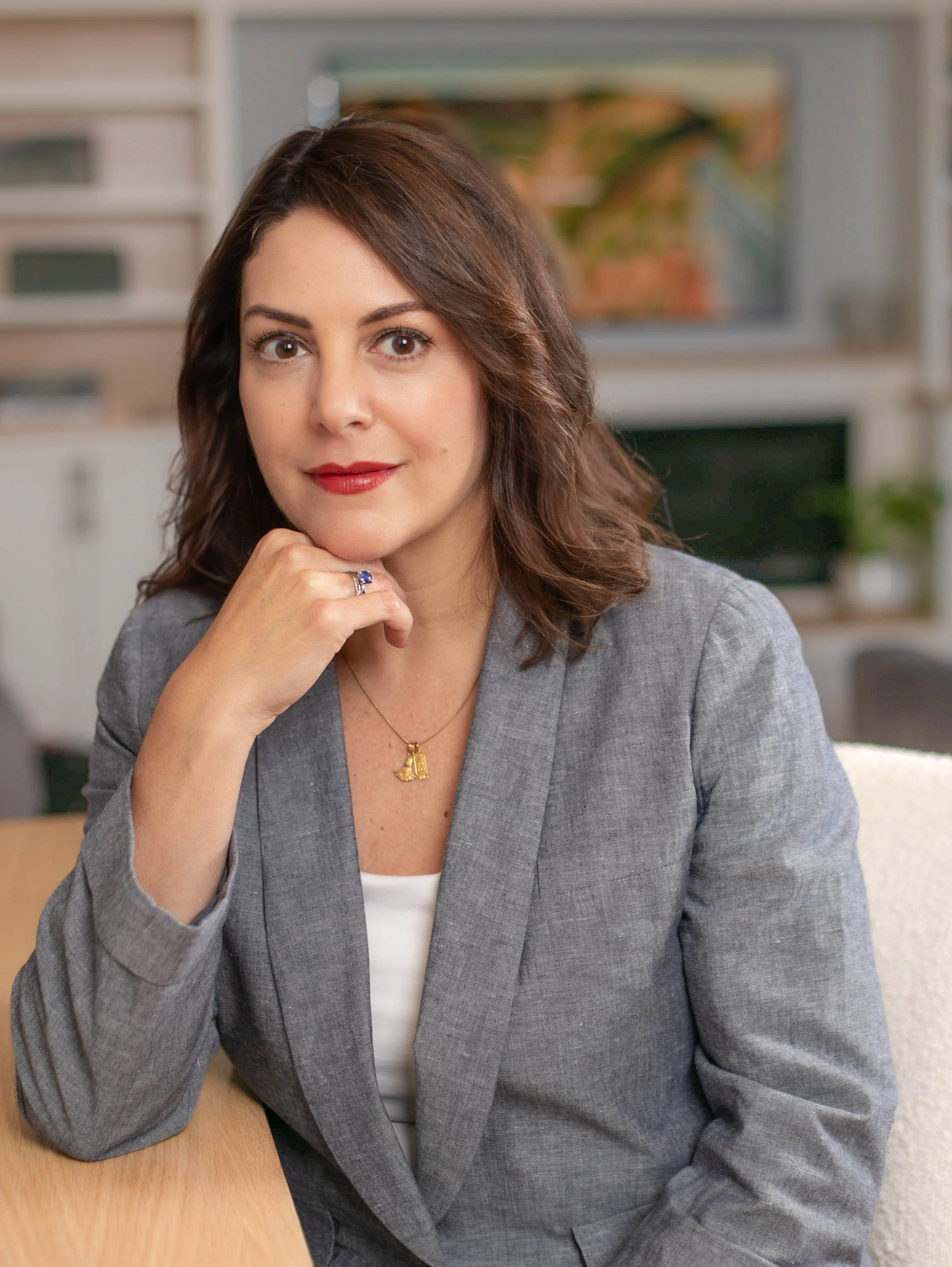 A woman with brown hair, wearing a gray blazer and white top, sitting at a table in an office or home setting, resting her chin on her hand, looking at the camera.