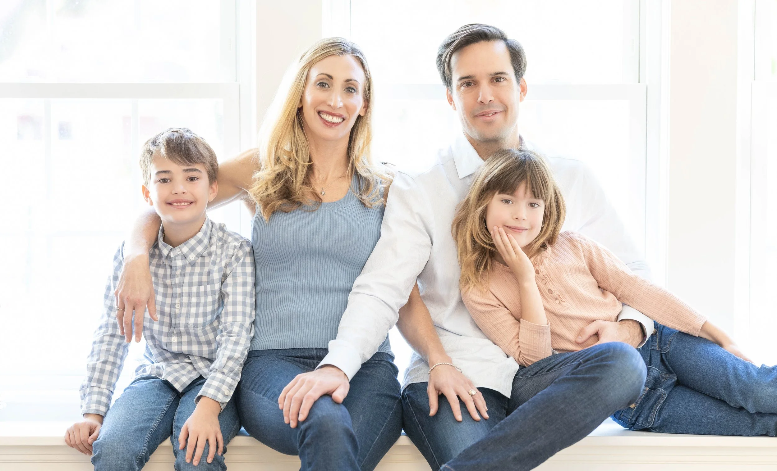 A family of four sitting on a white windowsill near a bright window, smiling at the camera