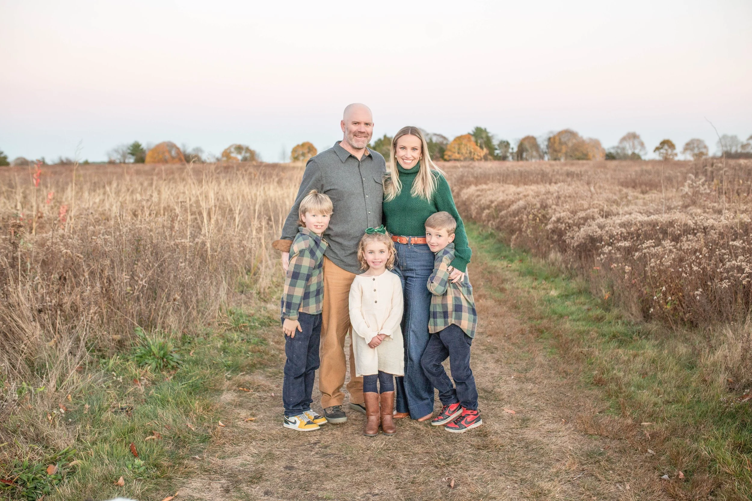 Family of five outdoors standing on a dirt path surrounded by tall grass and autumn trees, smiling at the camera.