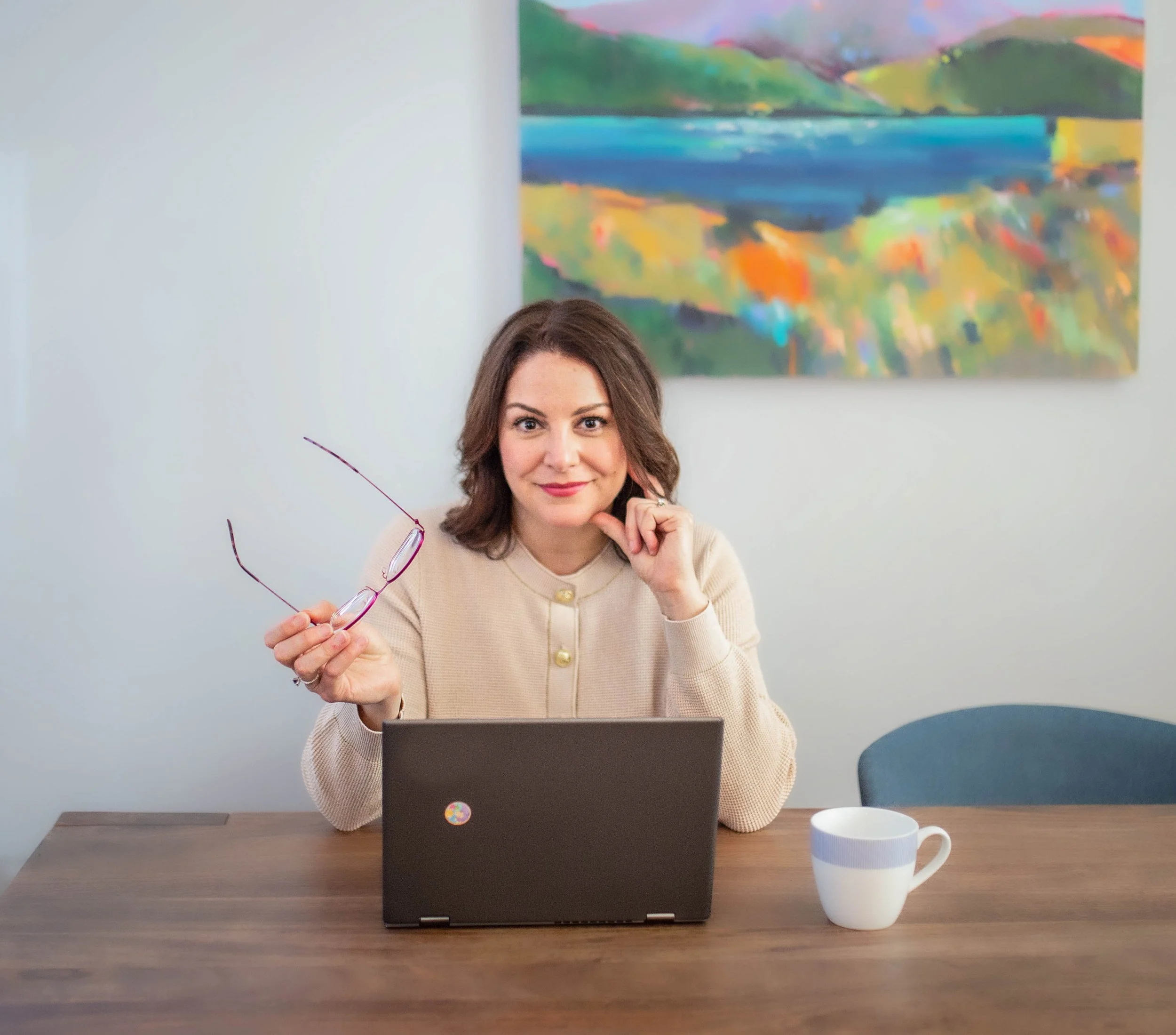 A woman with shoulder-length brown hair and a light beige sweater sits at a wooden table with a laptop, holding a pair of eyeglasses in her right hand and resting her chin on her left hand. There is a white mug with blue and gray stripes on the table