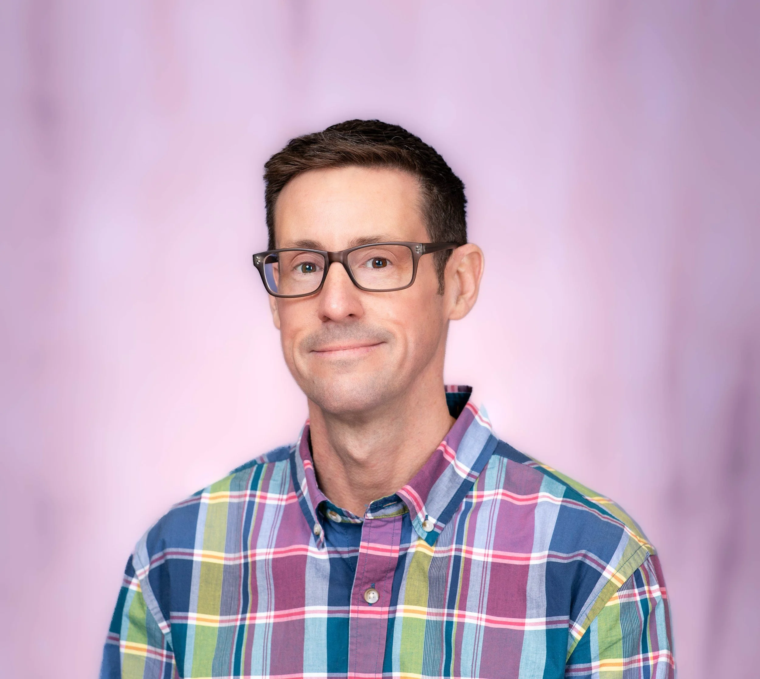 A man wearing glasses and a colorful plaid button-up shirt. Background is blurred with pink and purple tones.