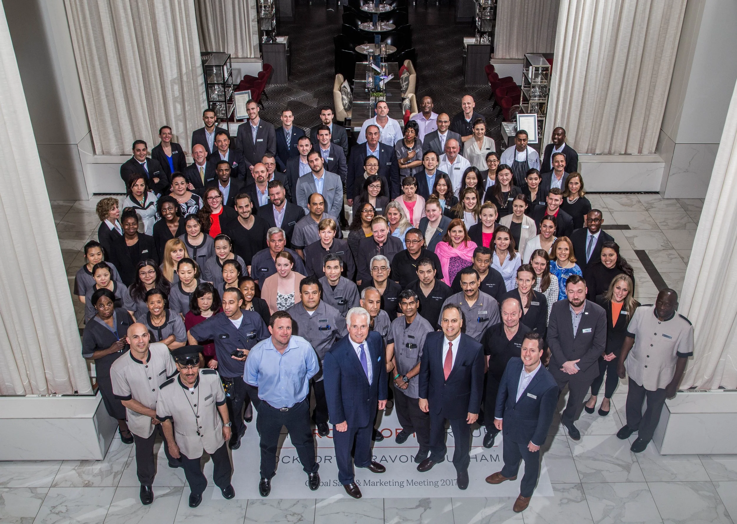 A large group of diverse people gathered in a hotel lobby for a corporate event, posing for a group photo from an overhead perspective.