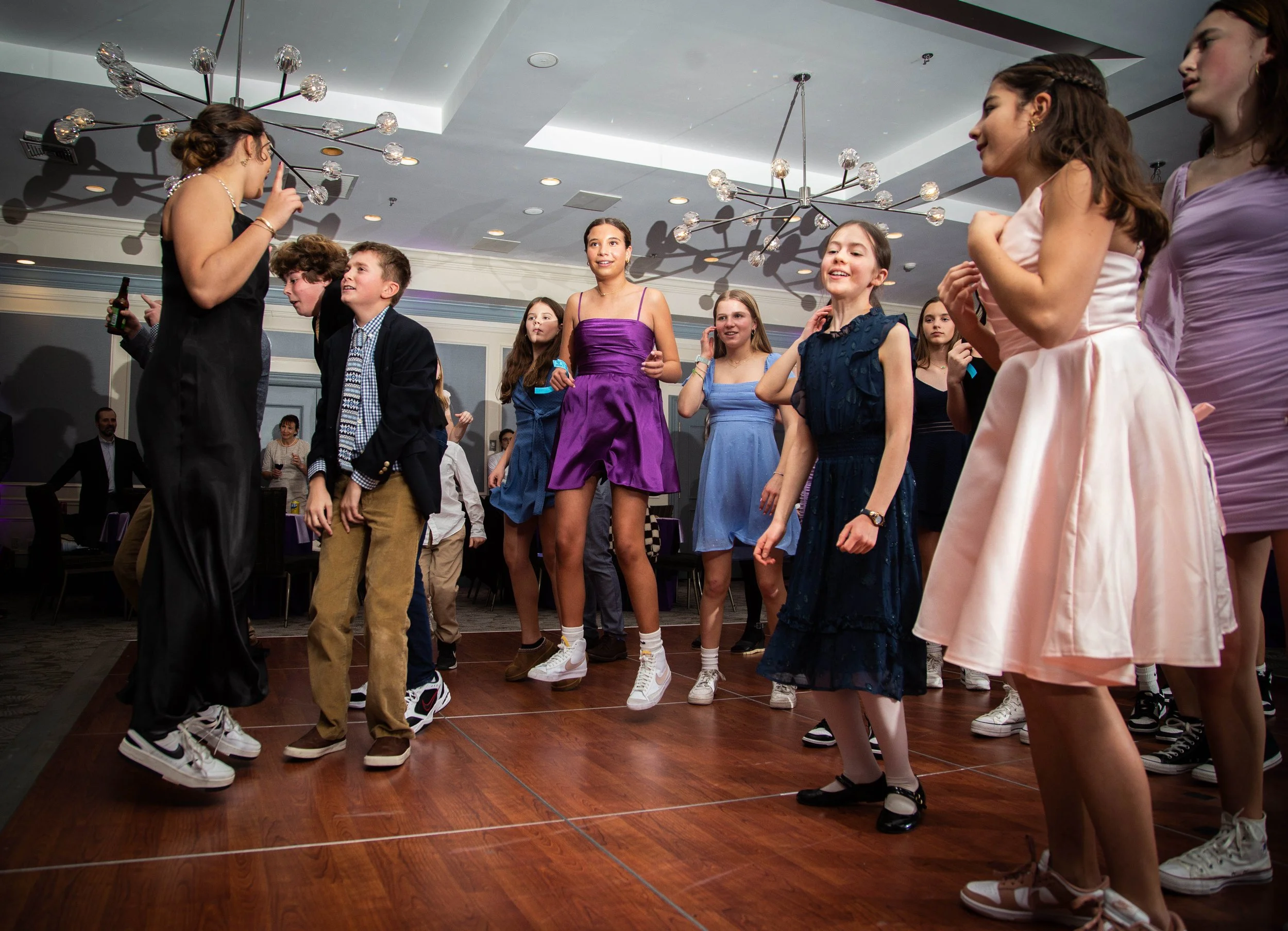 A group of young teenagers dancing and having fun at a party or social event in a banquet hall, with a woman hosting or DJing at the front.