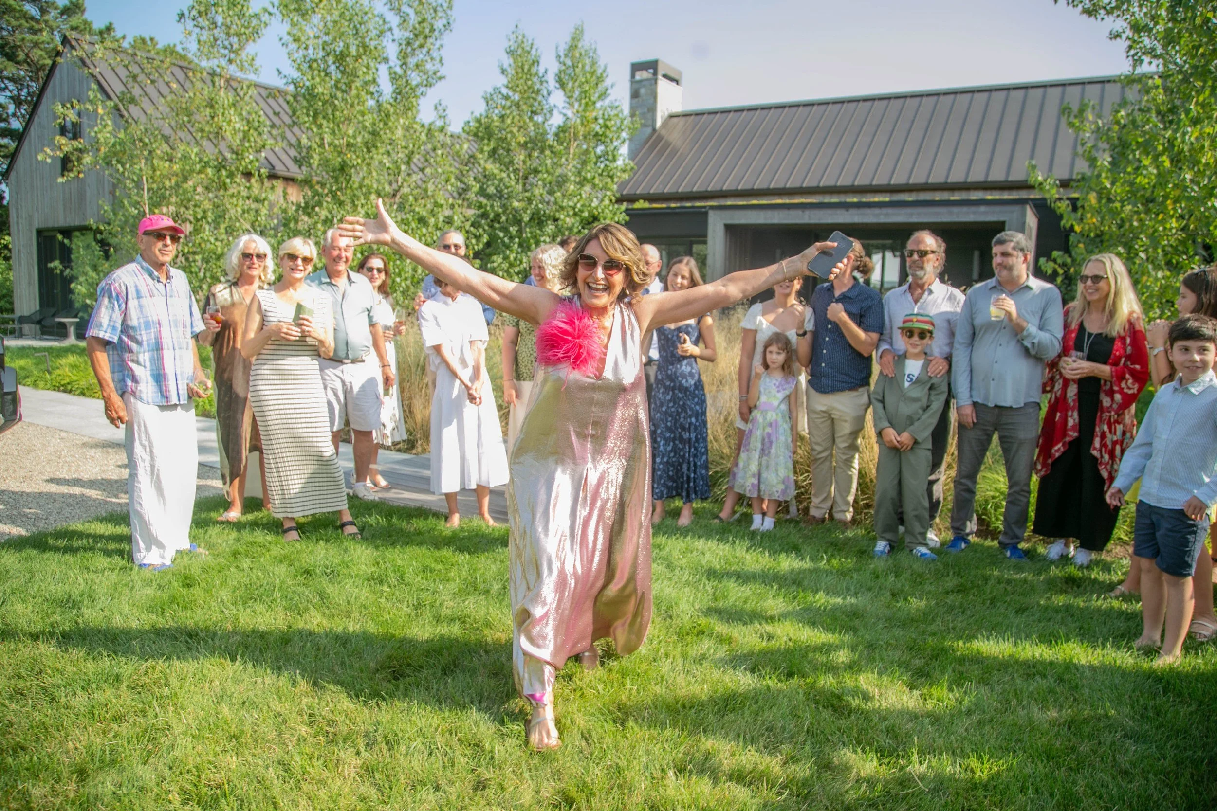 A woman in a shiny gold dress with a pink feather on her shoulder stands with arms outstretched, smiling broadly in front of a group of people at an outdoor event on a sunny day.