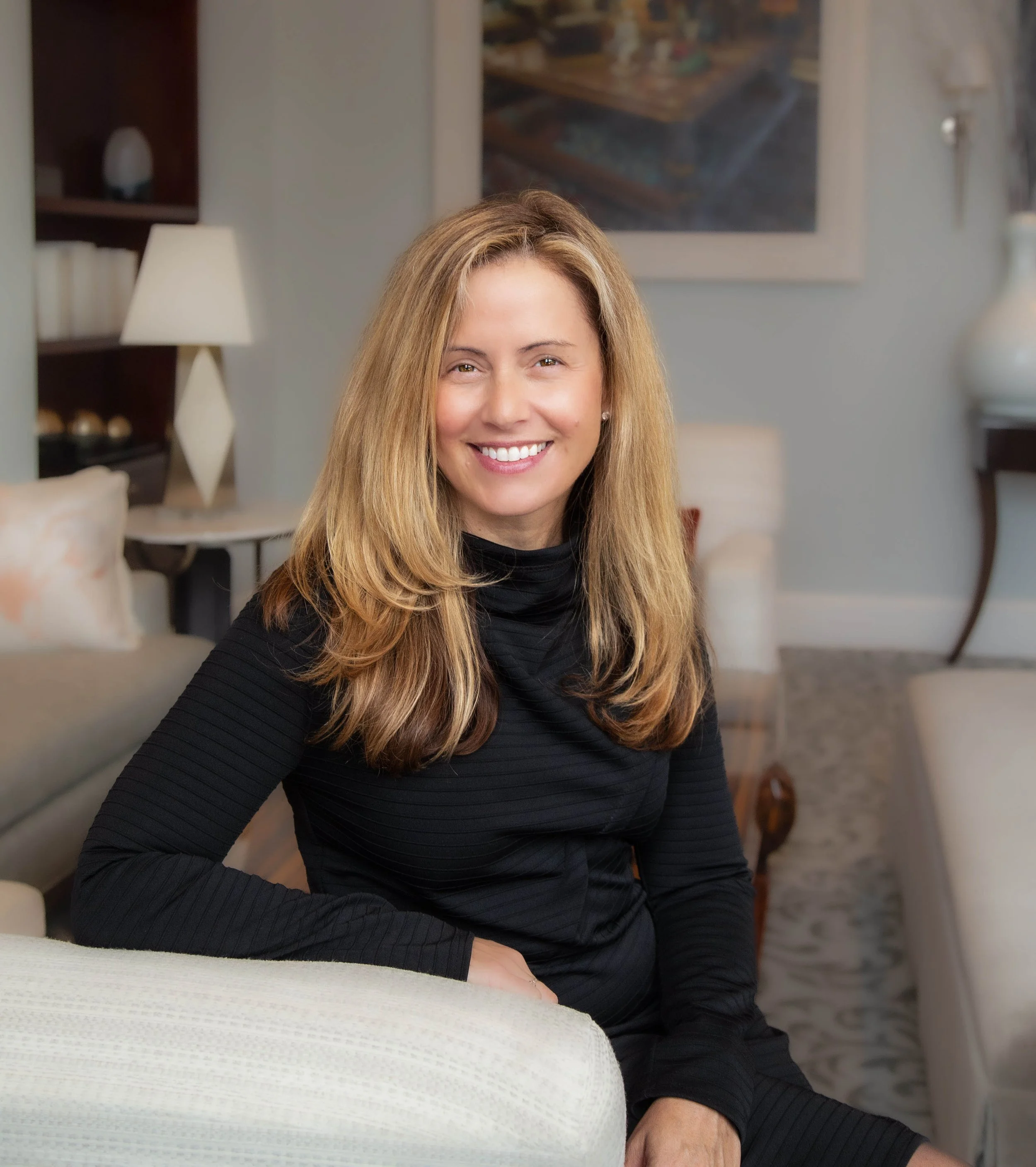 A woman with blonde hair smiling, sitting on a light-colored armchair in a cozy living room with art and lamps in the background.