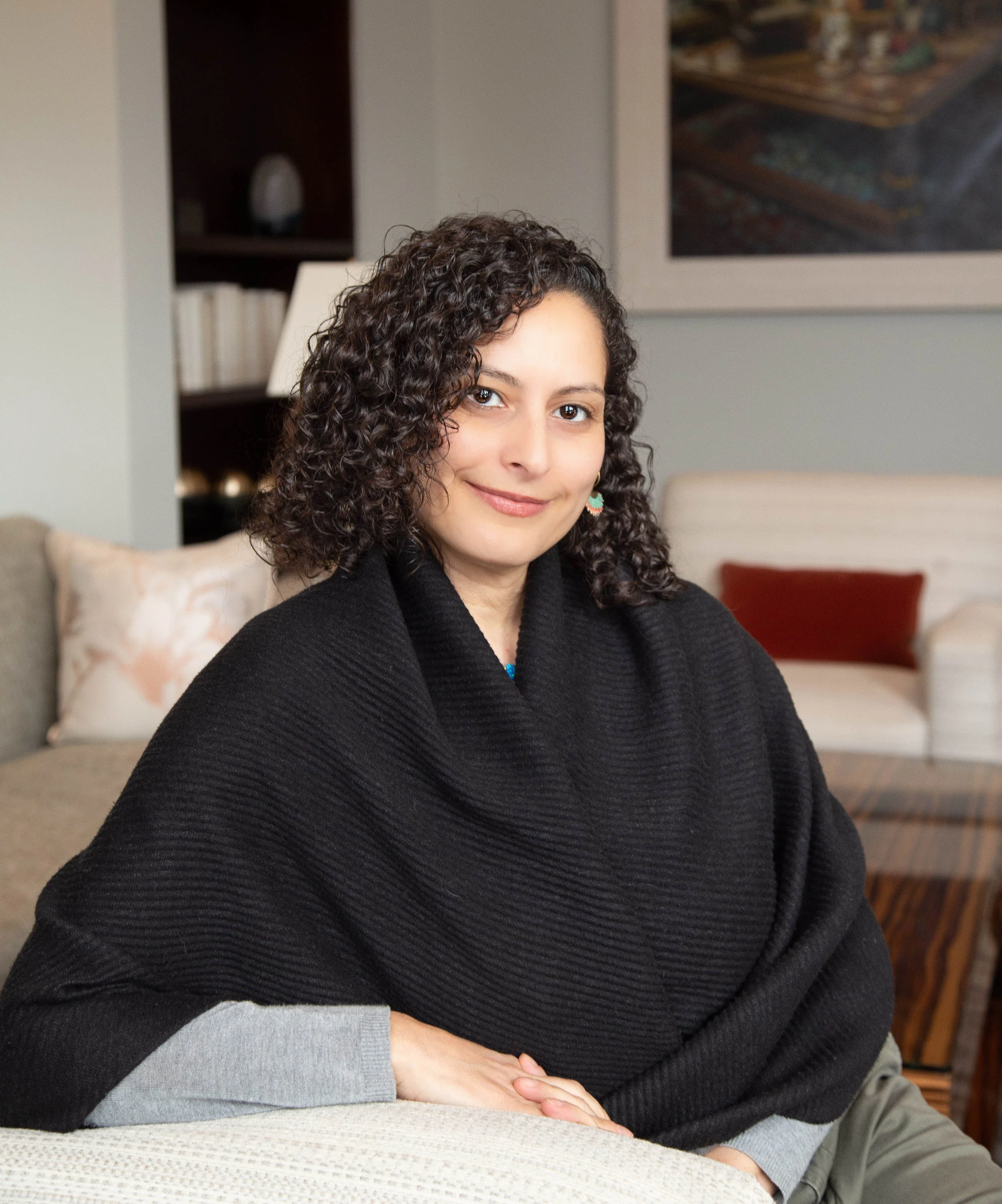 A woman with curly dark hair sitting on a sofa in a living room, smiling at the camera, wearing a black shawl over a gray long-sleeve shirt.