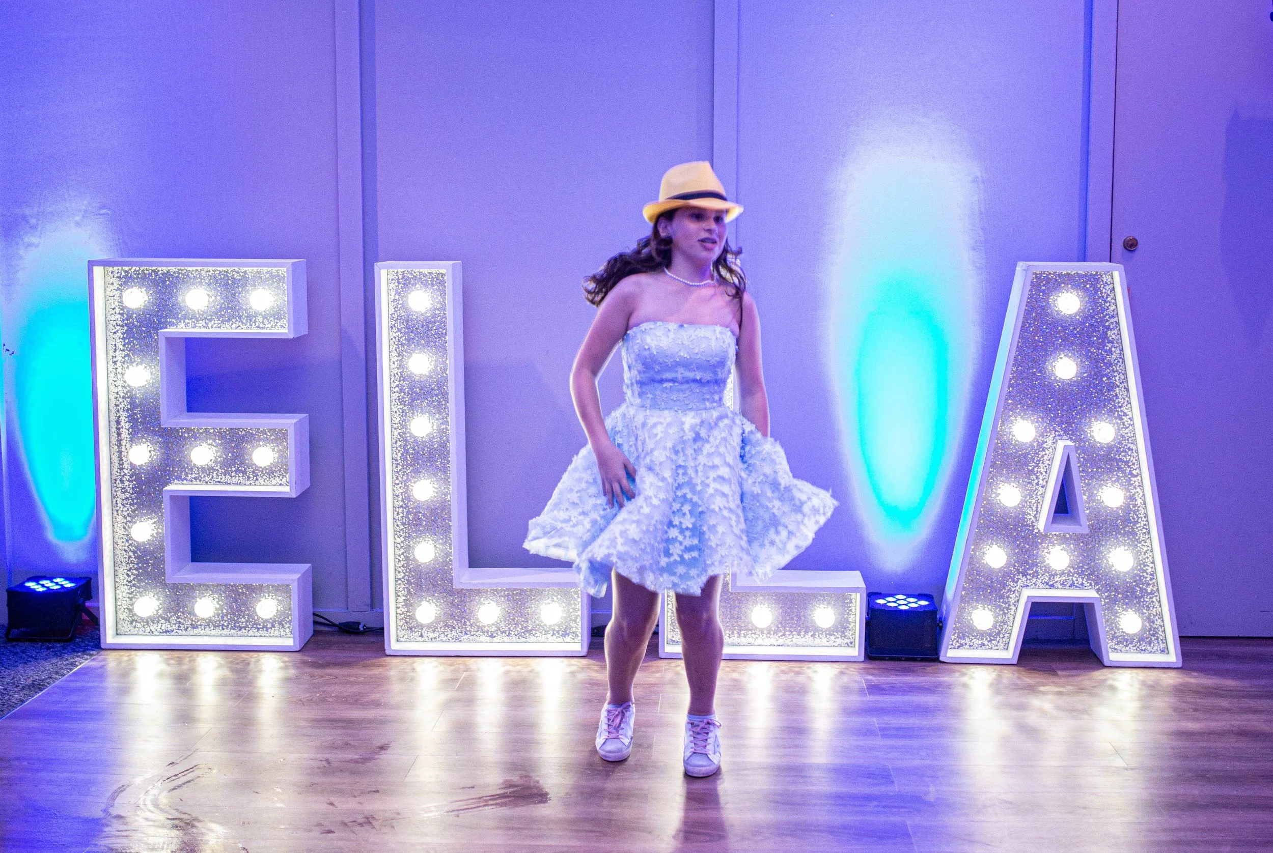 A young girl in a white dress and hat is standing in front of large illuminated letters spelling 'ELLA' at a party or event, with a wooden floor and purple-blue lighting in the background.