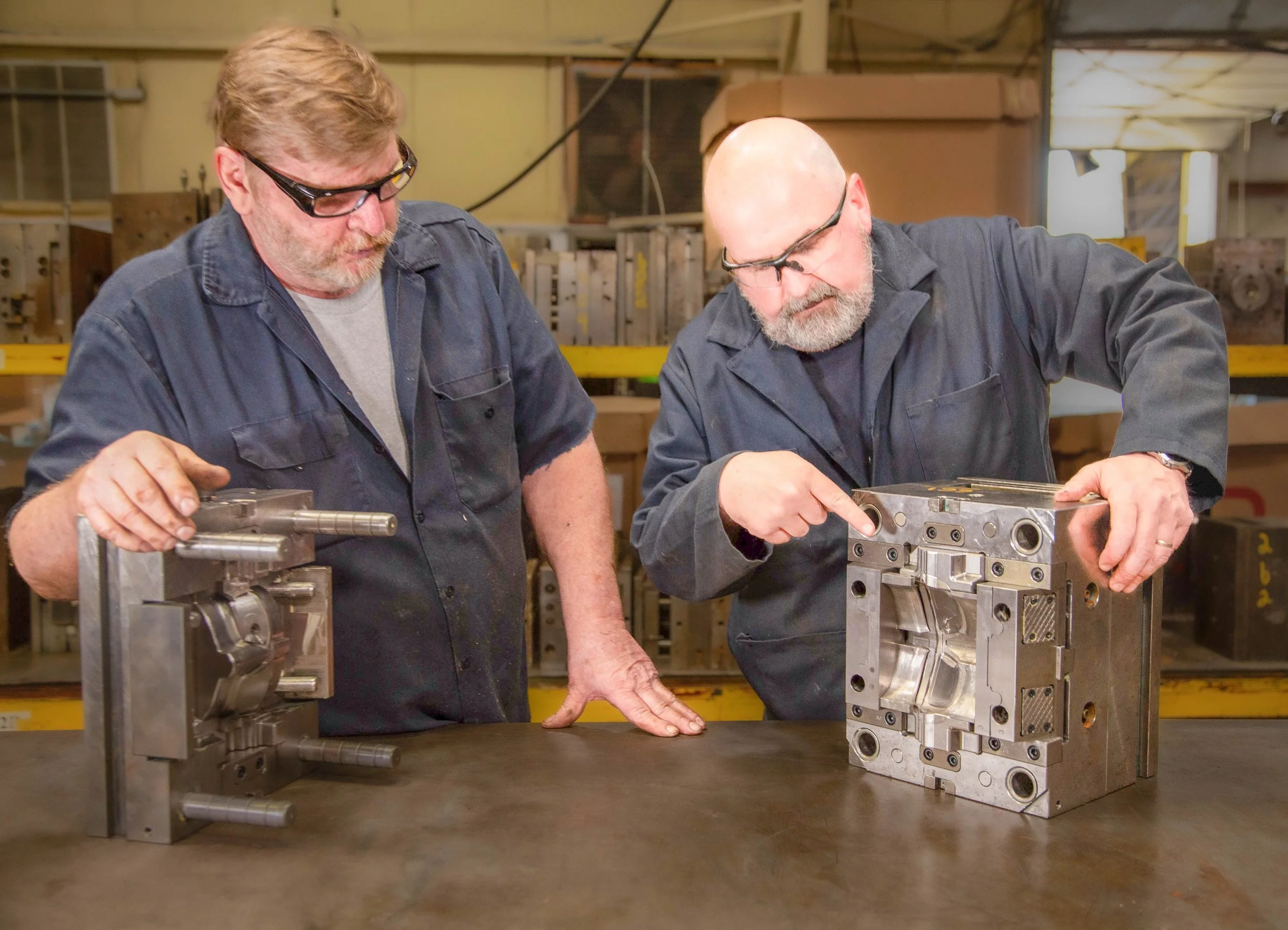 Two men wearing safety glasses and dark work uniforms examining a metal mold or machine component in a workshop or manufacturing setting.