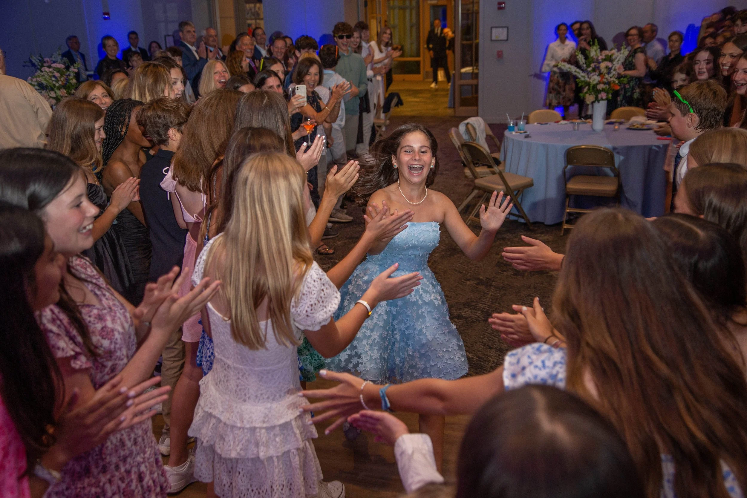 A young woman in a blue strapless dress smiling and dancing at a celebration, surrounded by a group of people clapping and cheering.
